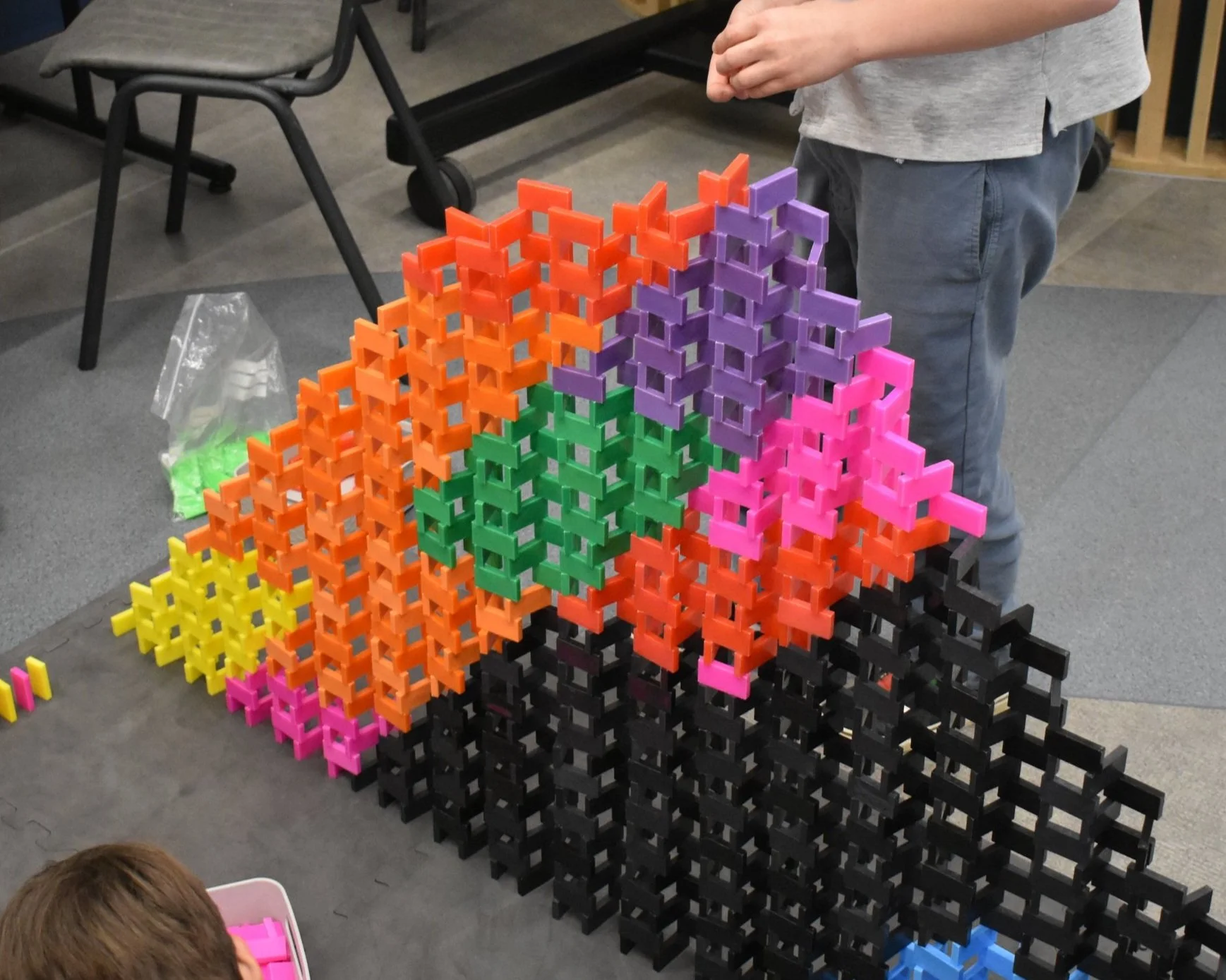 A 2D domino pyramid built in an array of colours, with a person standing nearby and a child with a pink container in front.