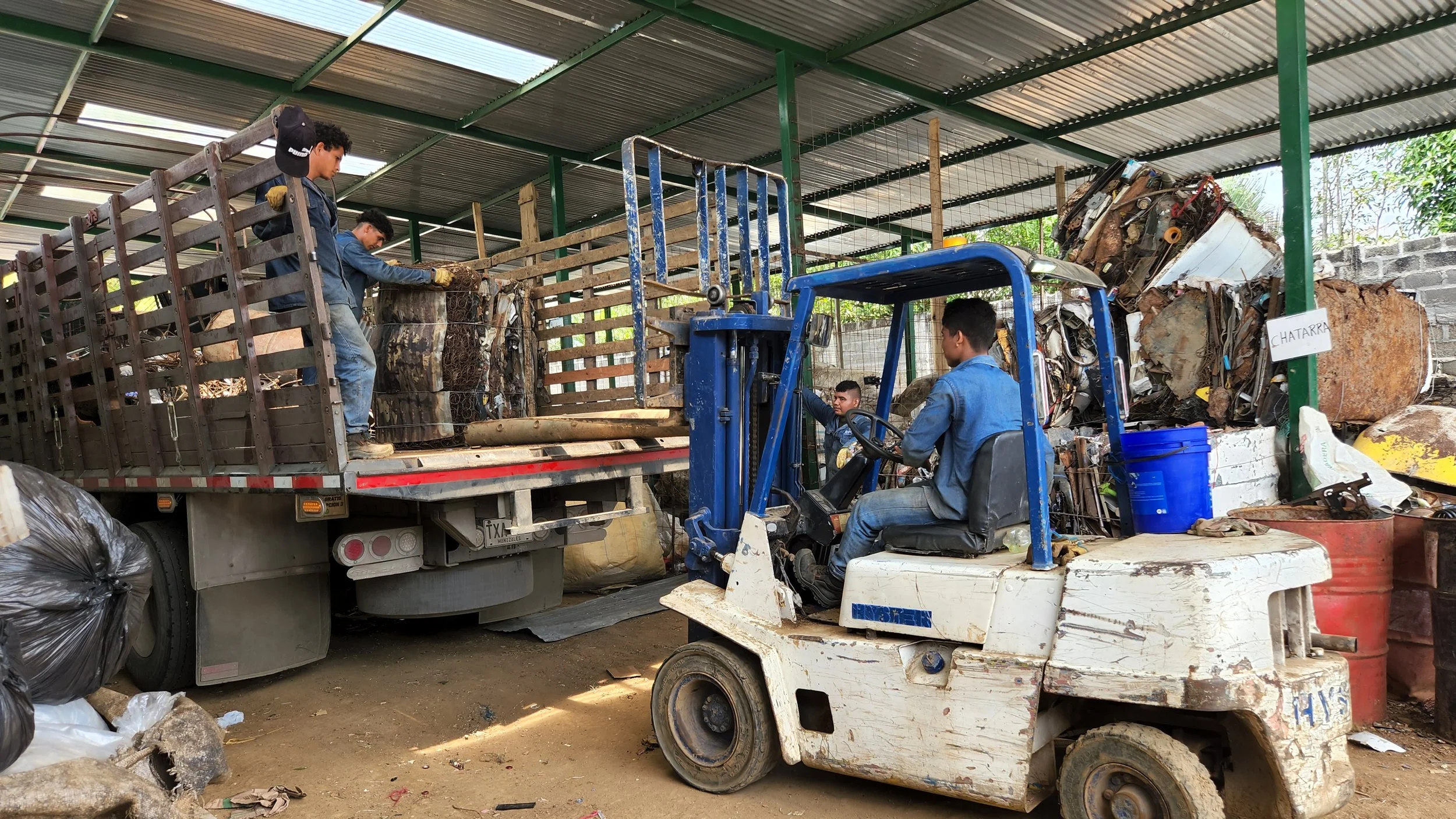Cuatro jóvenes trabajan en un centro de reciclaje, cargando y organizando materiales reciclables en un camión y una carretilla o montacargas