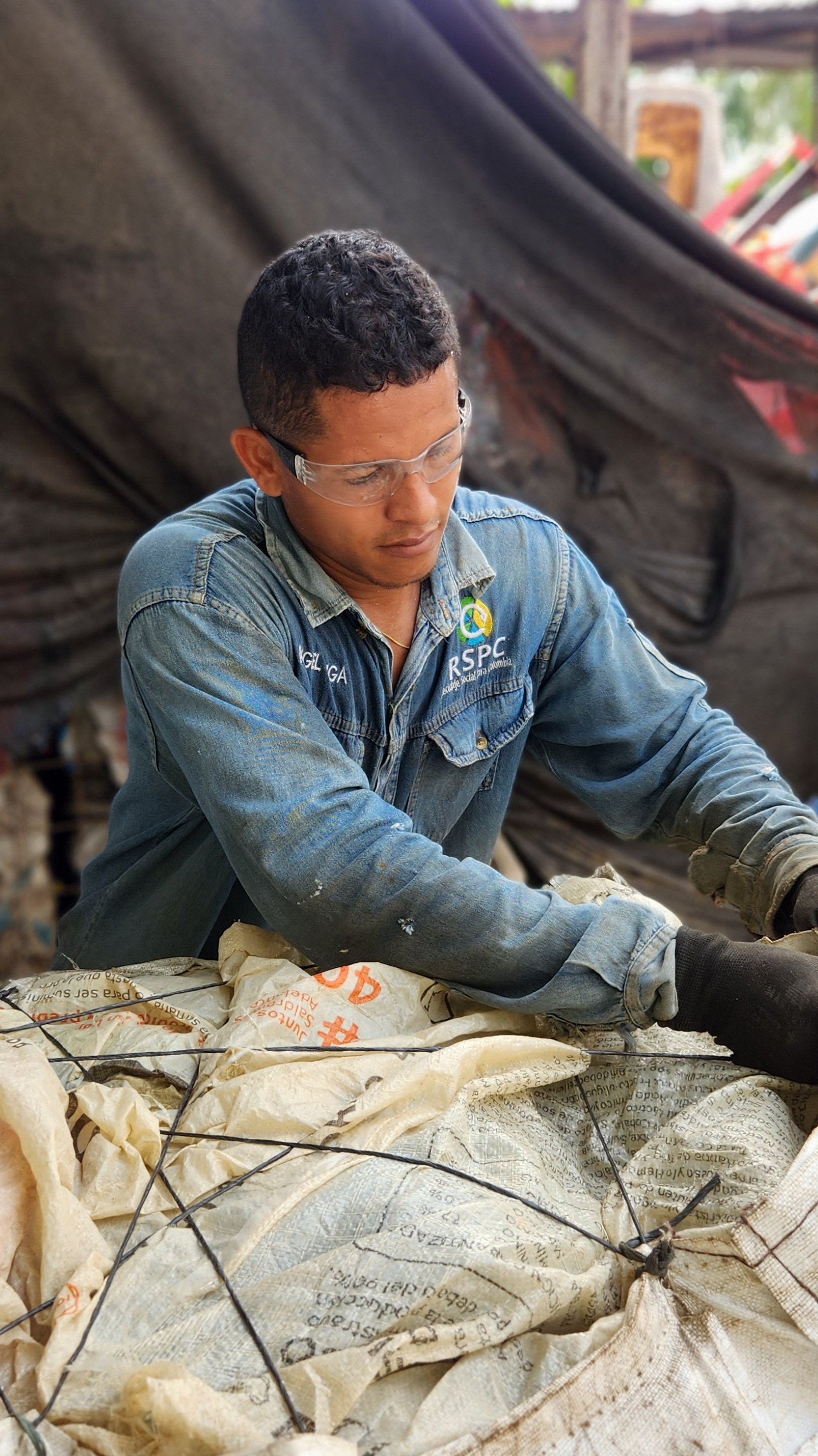 Un hombre con gafas de protección trabaja con sacos y varillas en un entorno al aire libre, posiblemente en un proyecto de construcción o reciclaje.