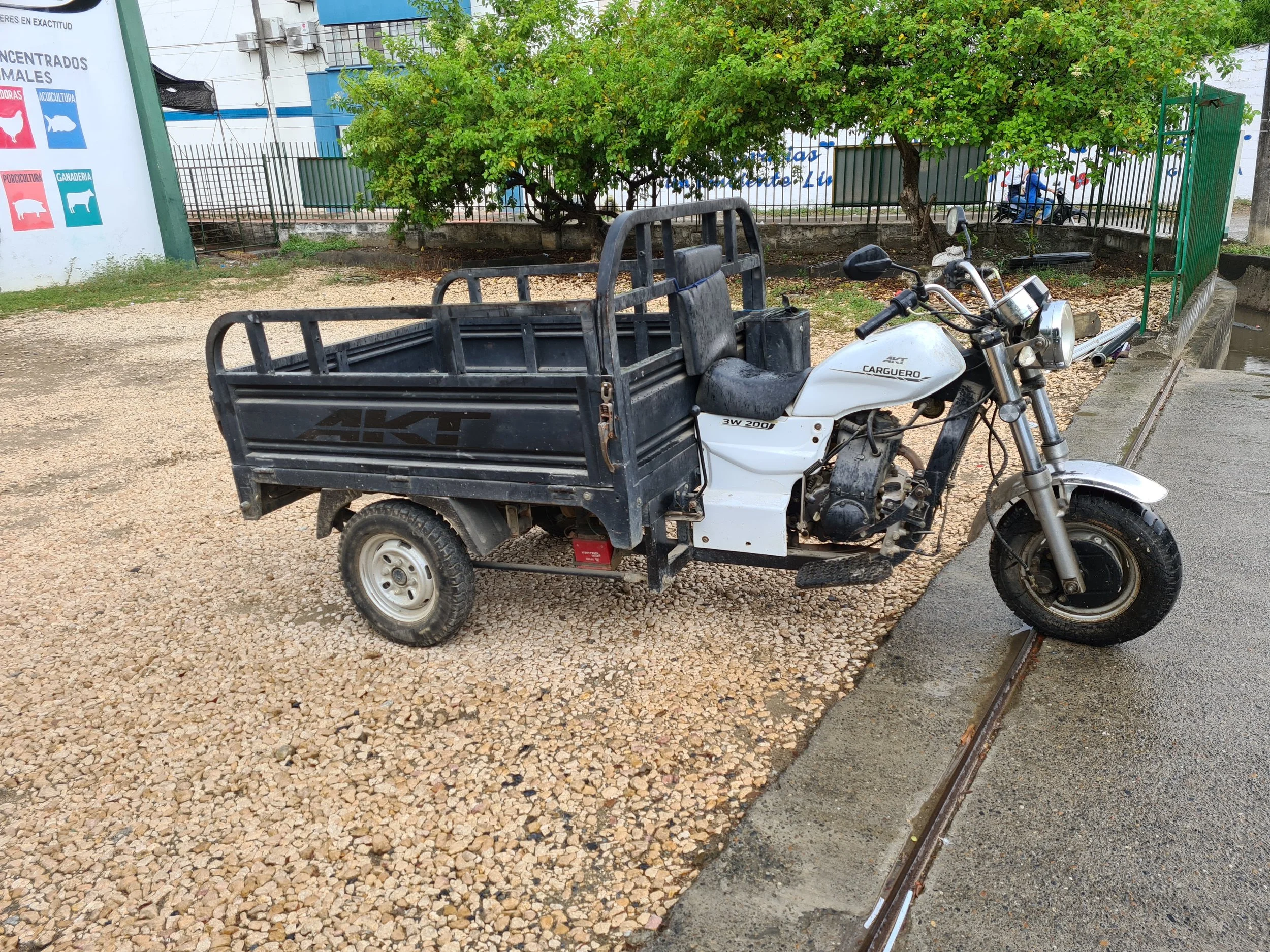 Una motocicleta de carga blanca y negra estacionada en un camino con grava, detrás hay árboles verdes, una cerca de metal y una pared con cartelera, en el fondo una persona monta bicicleta.