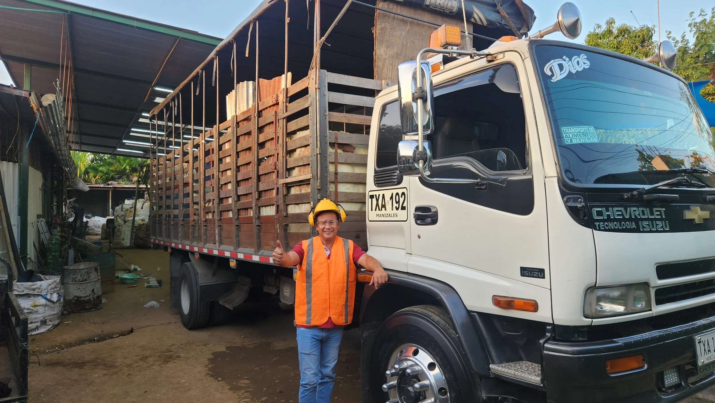Un hombre sonriente con casco y chaleco de seguridad, haciendo señal de pulgar hacia arriba, de pie junto a un camión de carga estacionado en un área de trabajo o taller, con estructura de techumbre y objetos en el fondo.