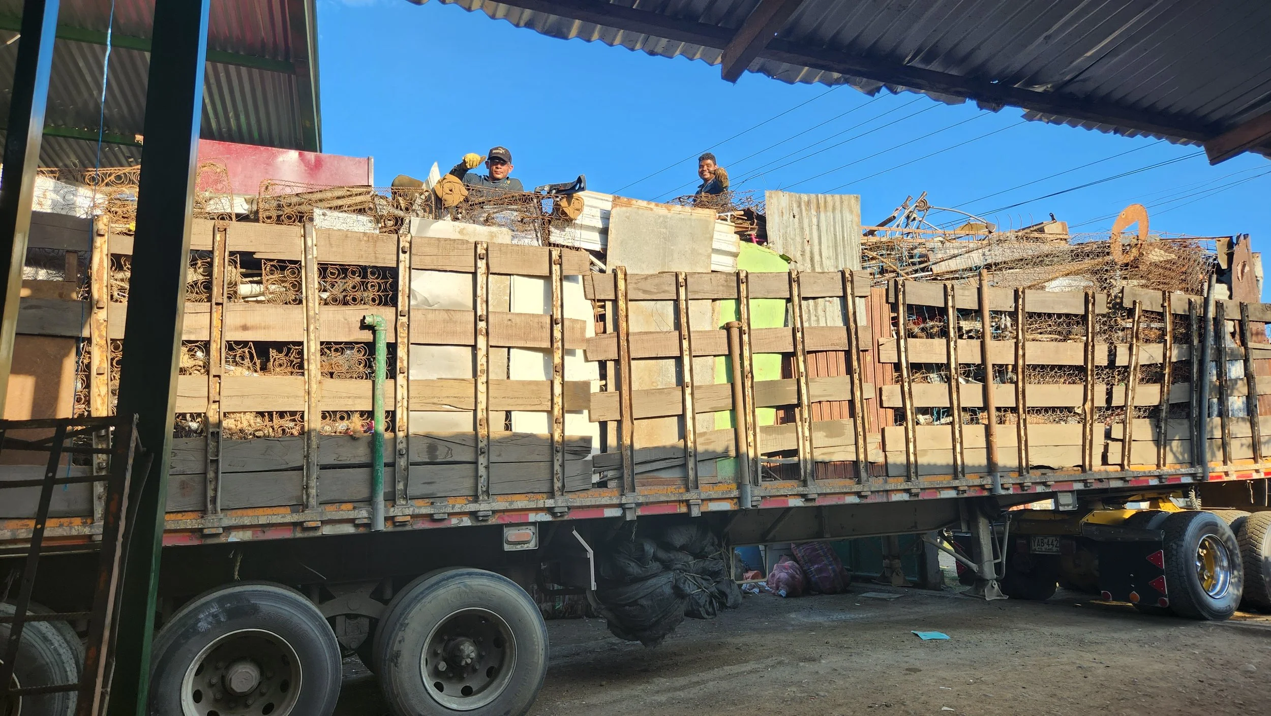 Dos hombres trabajando en la parte superior de un camión con carga de metal y materiales de construcción en un fondo despejado y cielo azul.