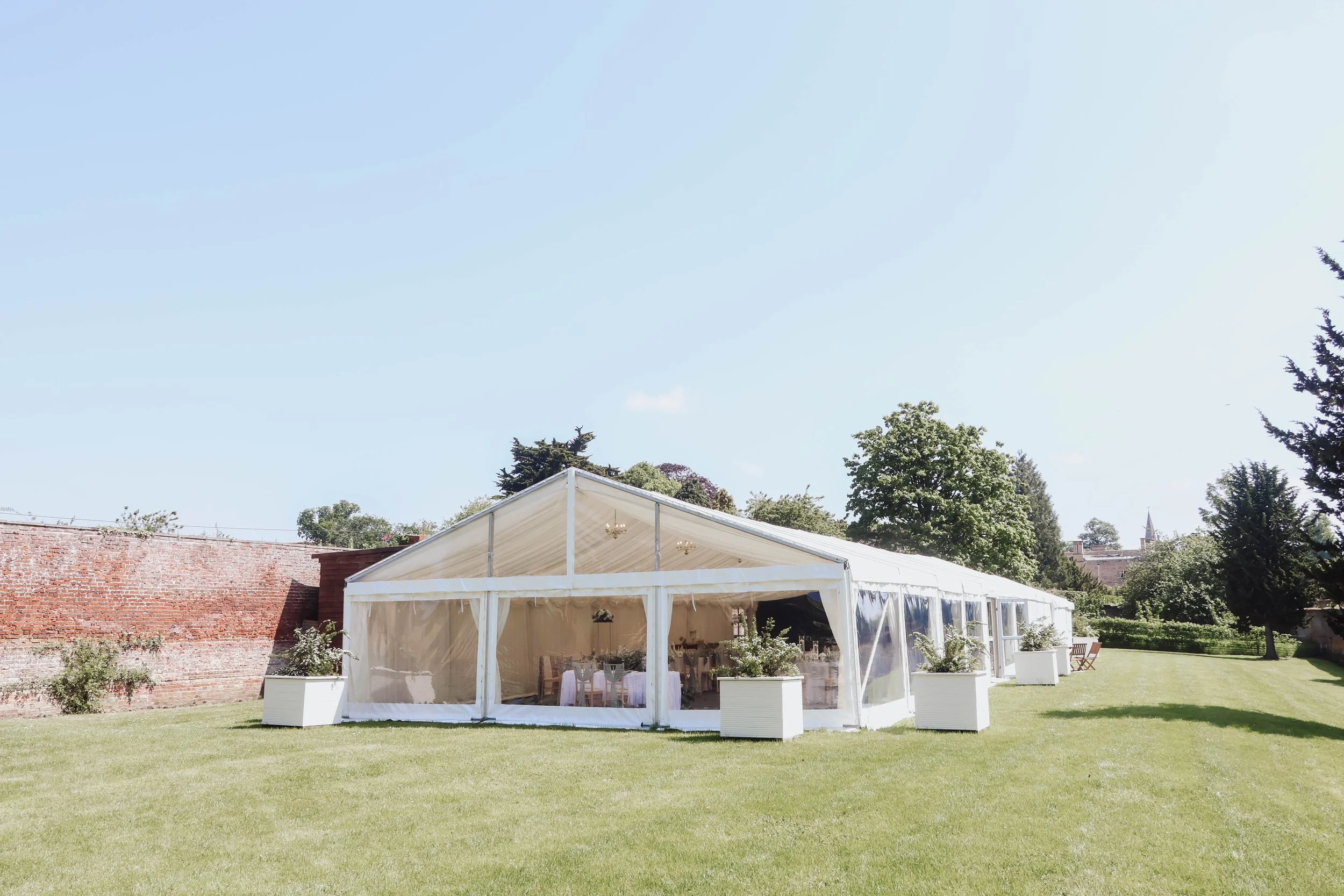 White event tent set up on a grassy area for a celebration, with tables and decorations inside, surrounded by trees and a brick wall, under a blue sky.