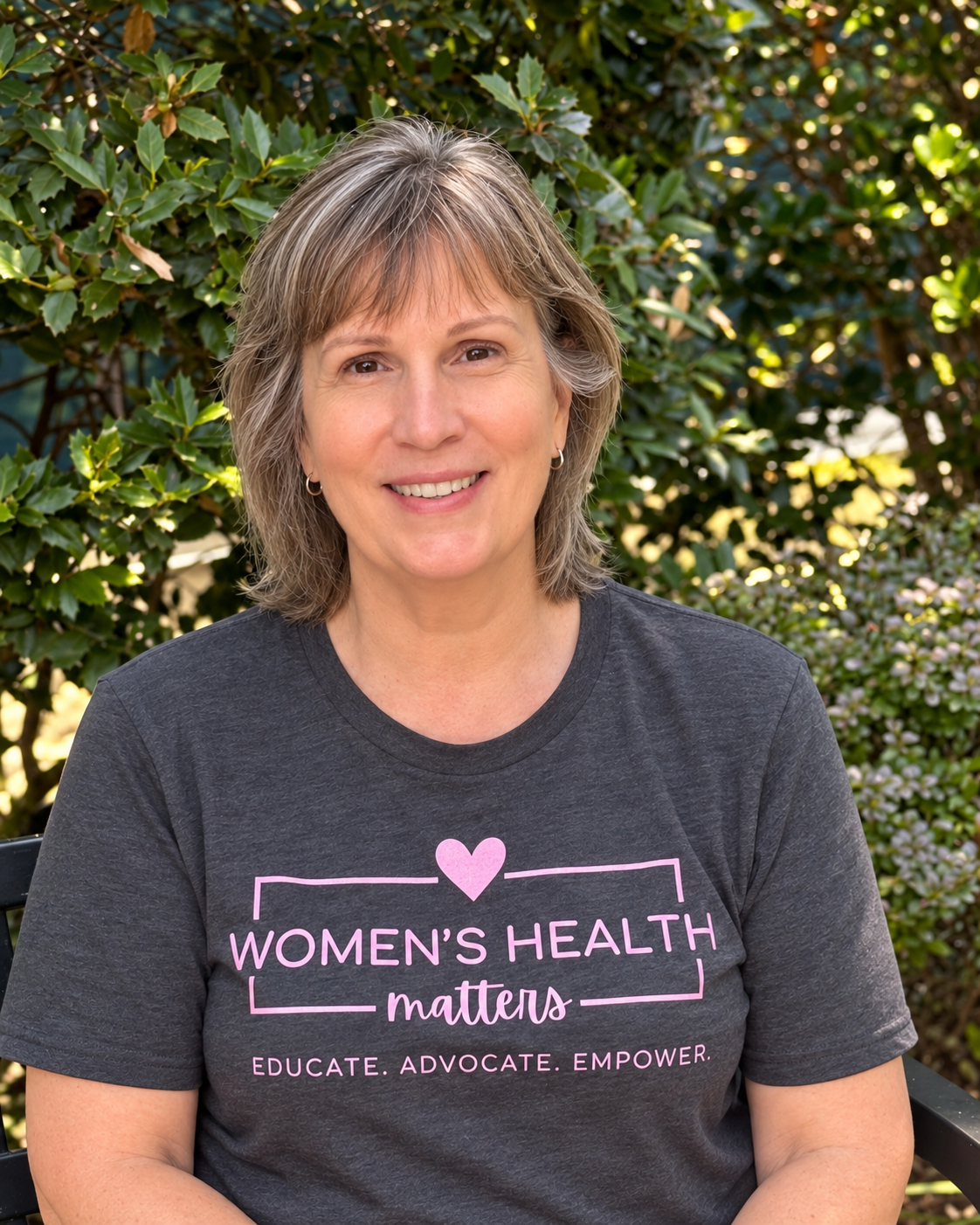 A woman with short, gray hair and hoop earrings, smiling outdoors in front of green foliage, wearing a gray t-shirt with pink text that reads 'Women's Health Matters,' along with a pink heart and the words 'Educate. Advocate. Empower.'