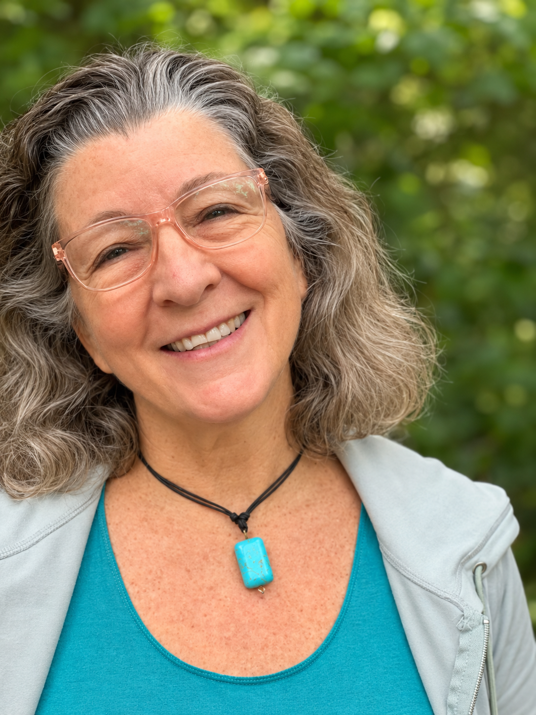 A smiling middle-aged woman with glasses, wavy gray hair, wearing a blue top, a gray jacket, and a turquoise necklace, outdoors with green foliage in the background.