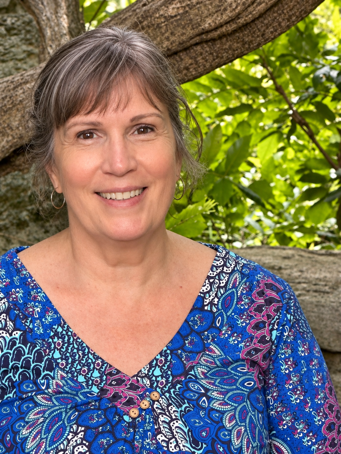 A smiling woman with short gray hair wearing a blue patterned top, standing outdoors near a tree with green leaves.