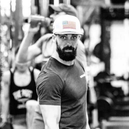 A man with a beard, wearing a cap with an American flag, in a gym.
