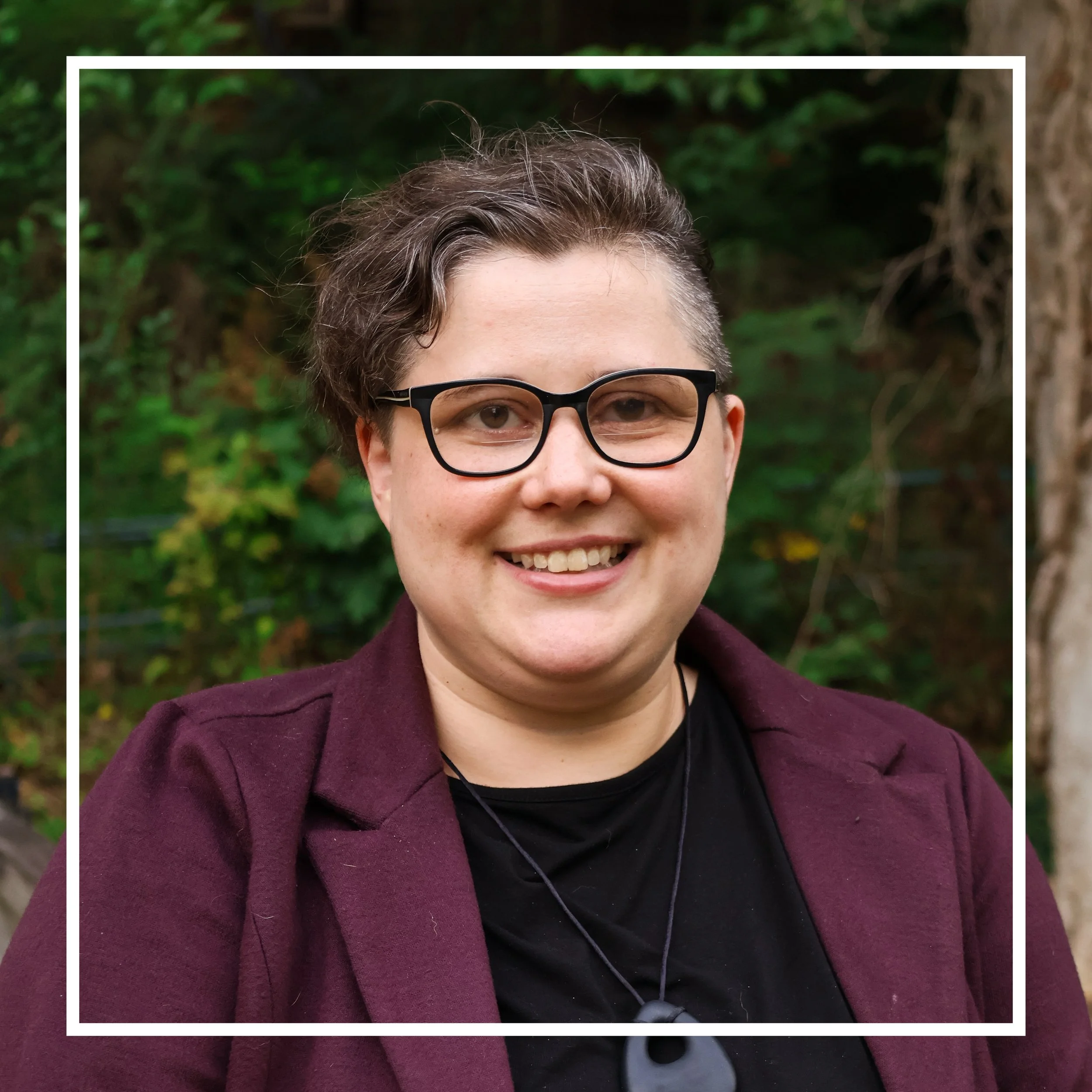 Close-up of a smiling woman with short hair, wearing black glasses, a dark shirt, a maroon coat, and earphones, standing outdoors near greenery.