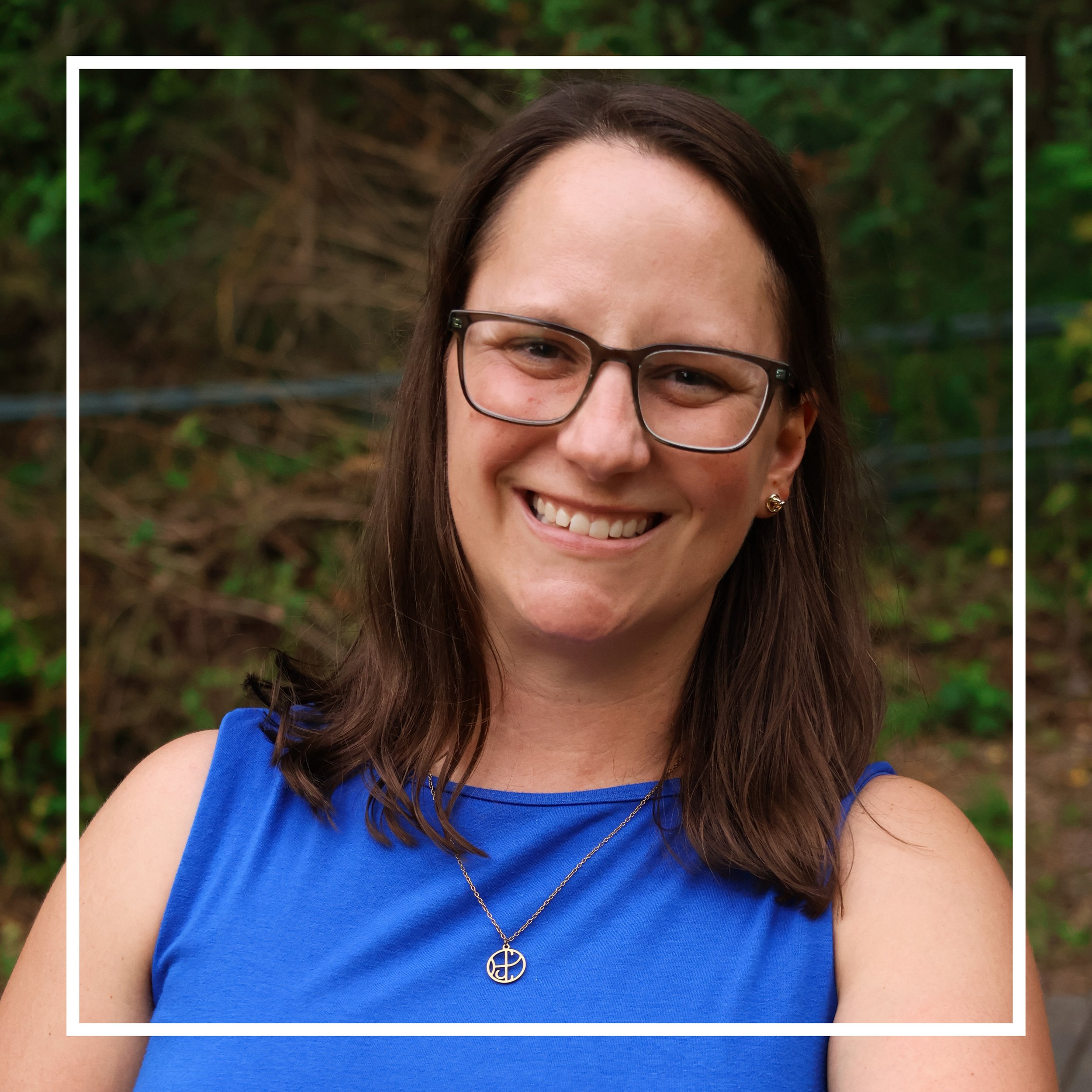 Brunette woman in blue shirt with glasses standing in front of greenery