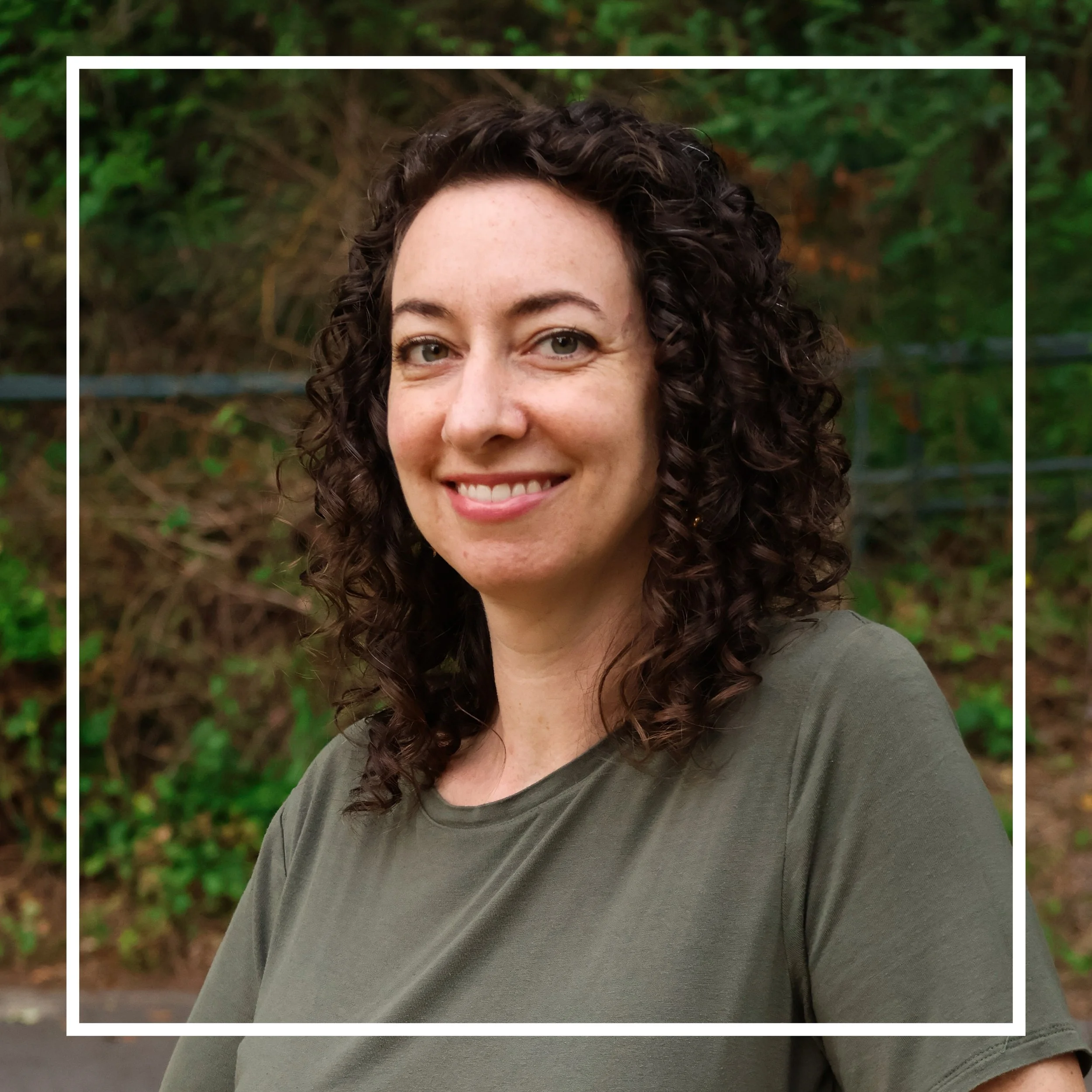 Close-up of a woman with dark curly hair smiling outdoors with green foliage in the background.