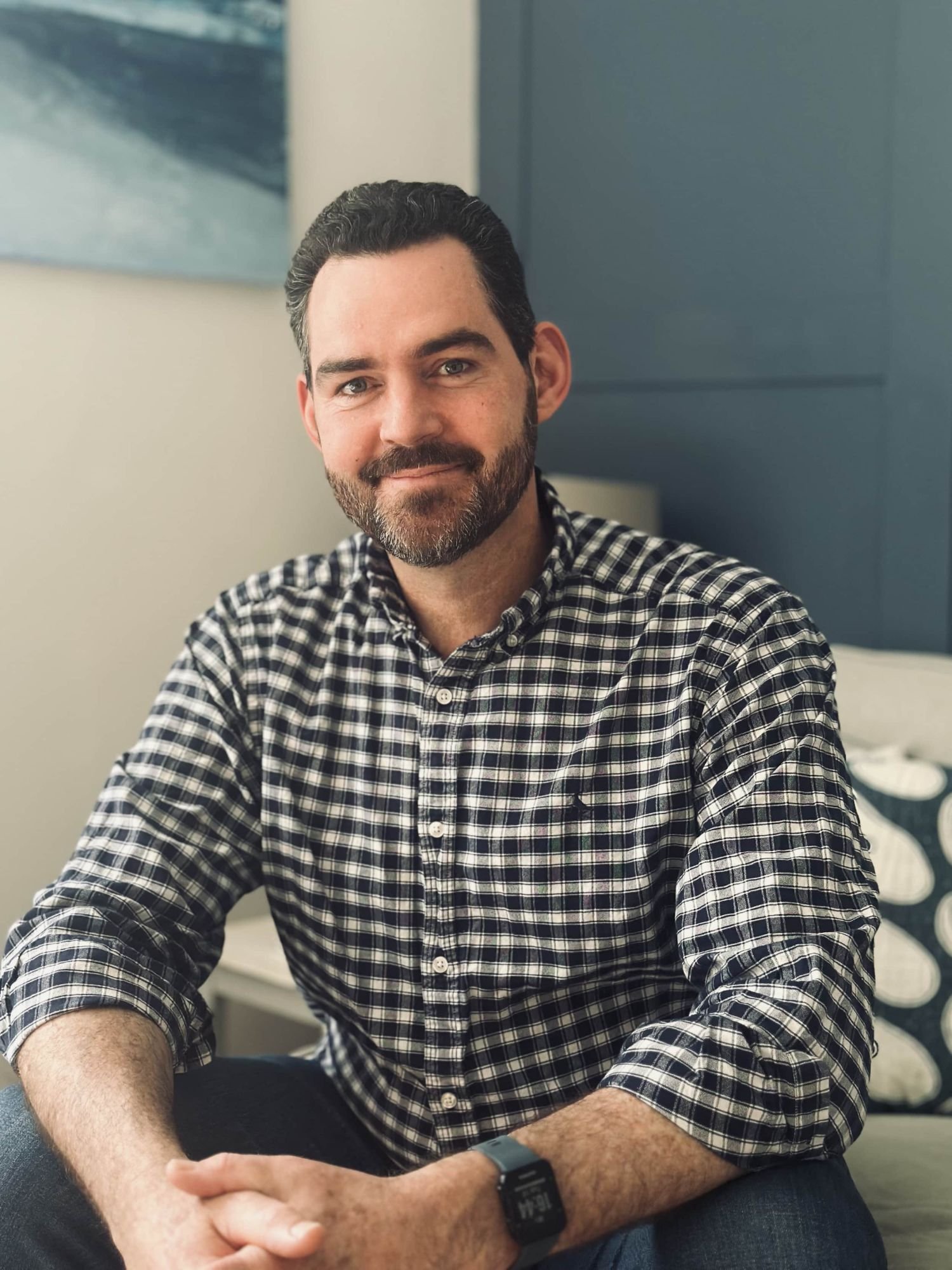 A man with a beard and dark hair, wearing a checkered shirt and a smartwatch, sitting indoors with a relaxed posture.