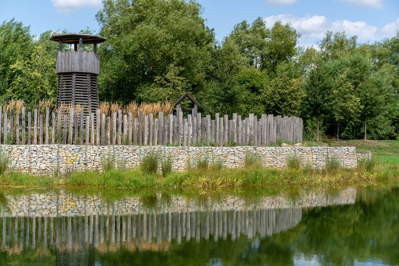 A wooden lookout tower, a small wooden structure, and a tall fence surrounded by trees and bushes near a body of water with a clear sky in the background.