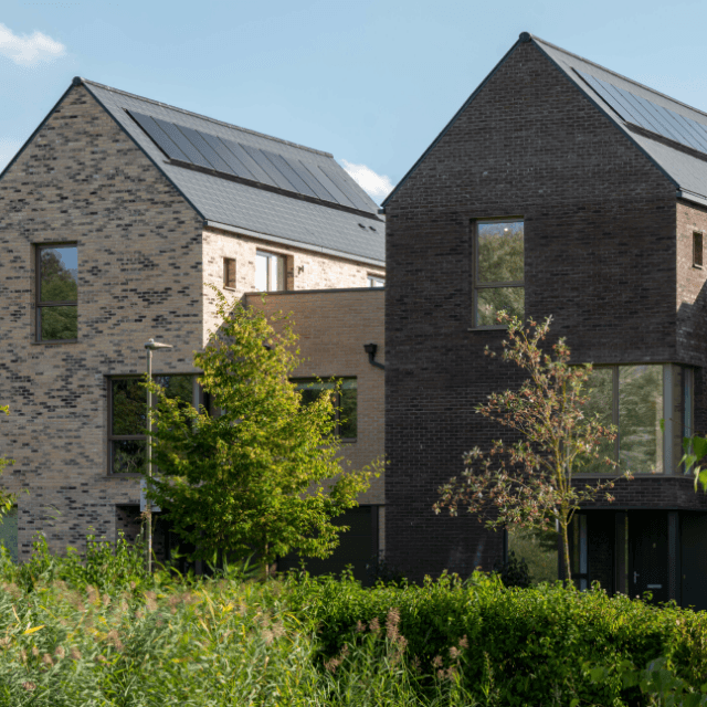 Two modern brick houses with solar panel roofs surrounded by trees and greenery.