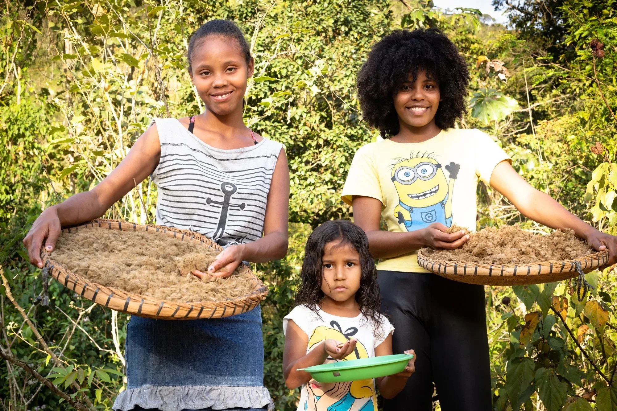Three girls standing outdoors holding woven trays with natural fibers, surrounded by vegetation