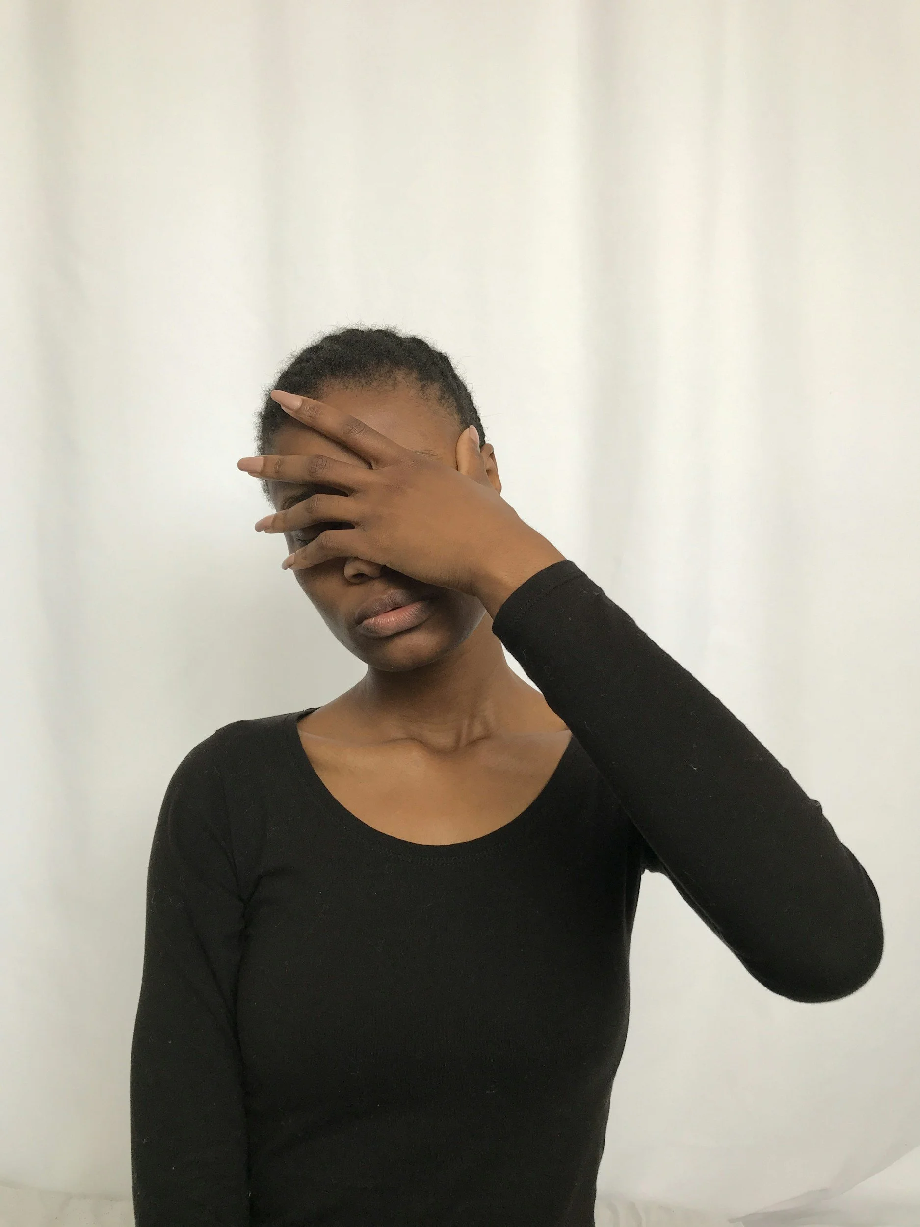 A young woman with short hair wearing a black long-sleeve shirt covering her face with her right hand against a plain white background.