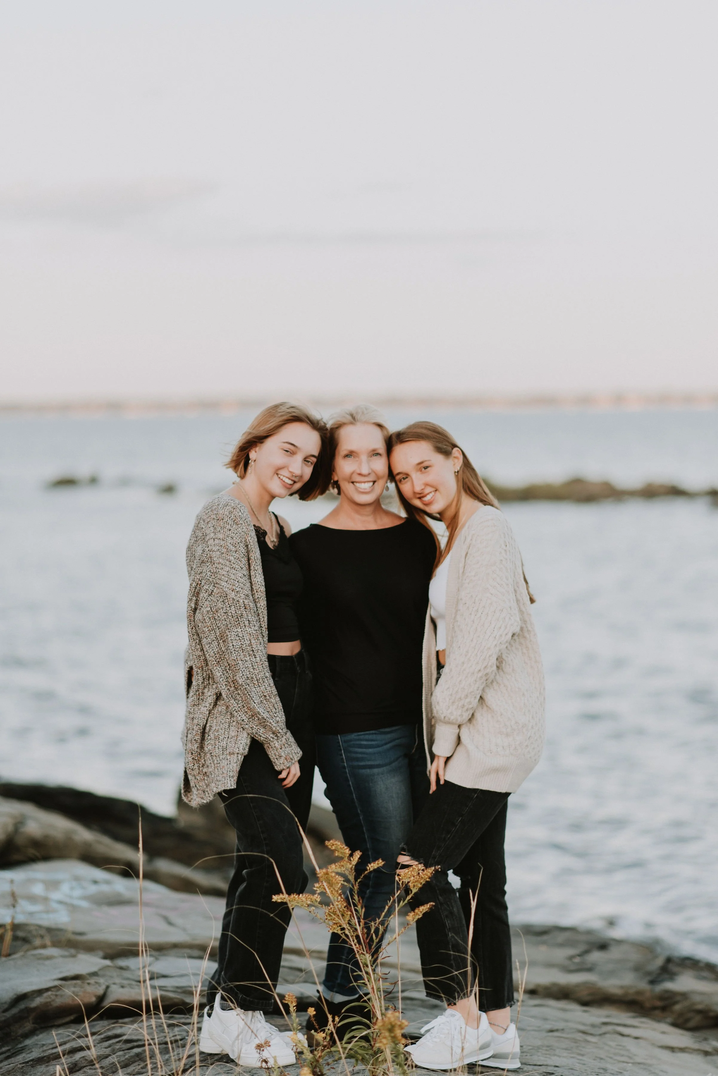 Three people smiling, standing on rocks by the water, wearing casual sweaters and jackets.