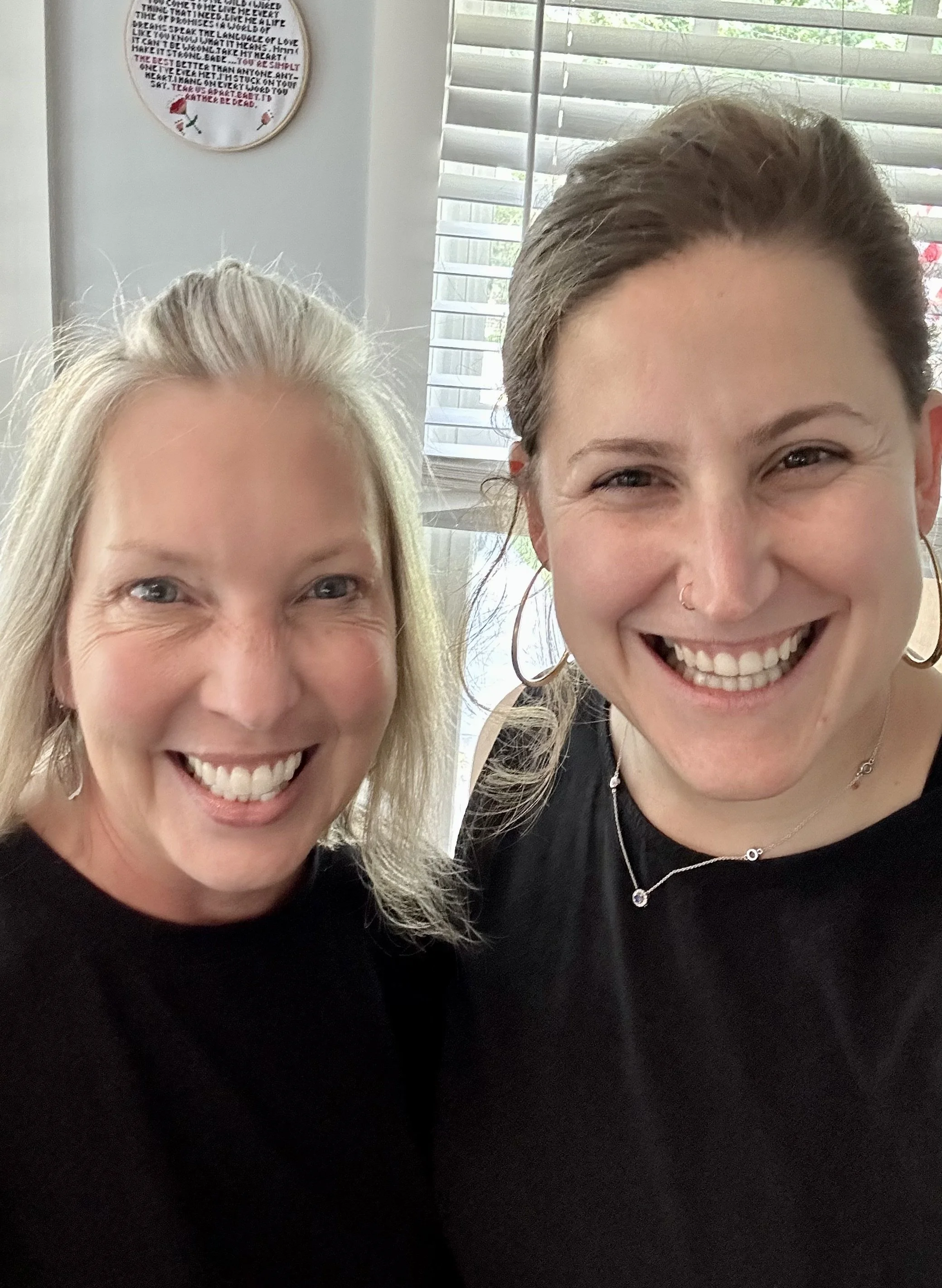 Two people smiling indoors, standing close together with a window and embroidery hoop in the background.