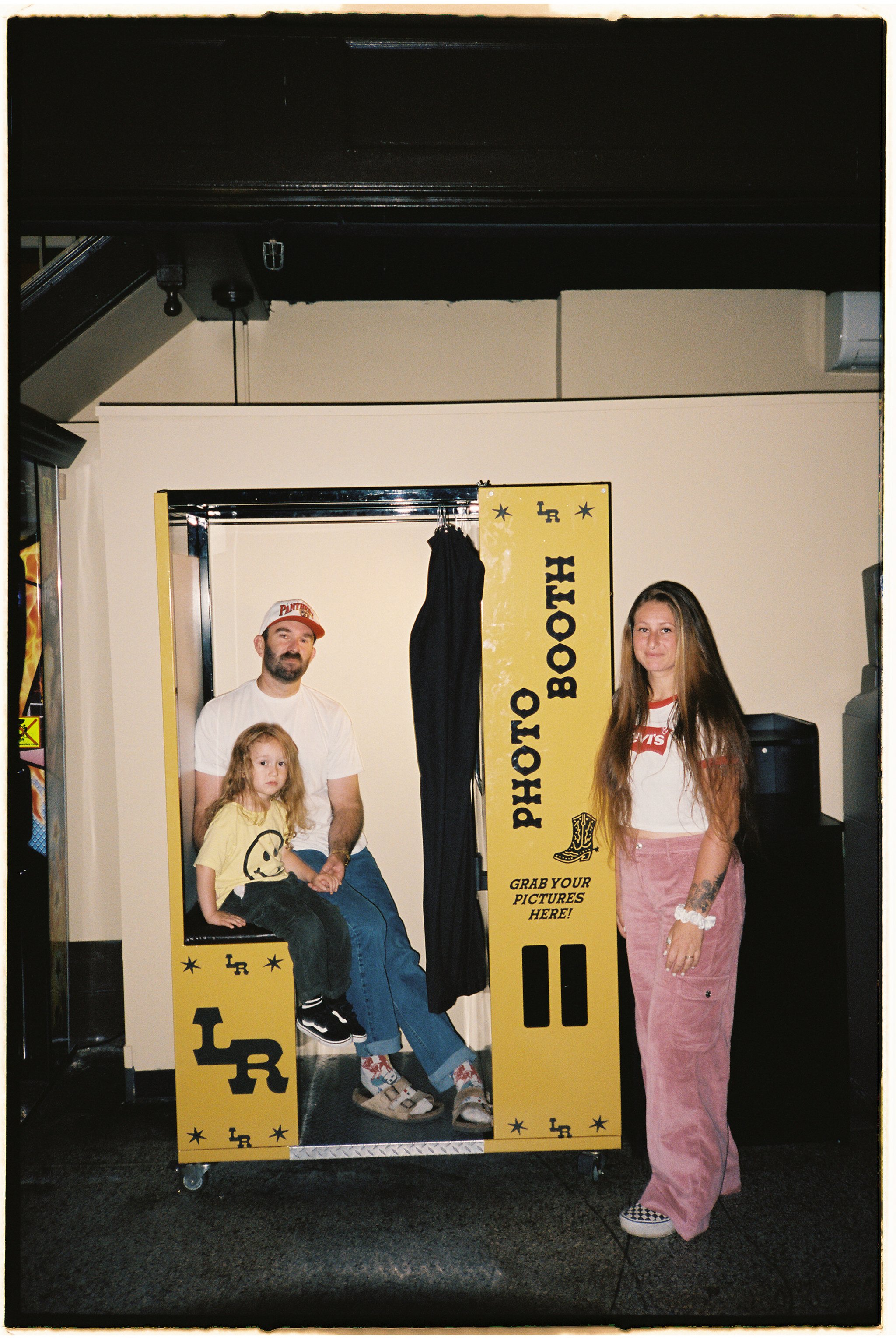 A family photo at a photo booth with a yellow backdrop and black curtain. The man is sitting on a seat with a child on his lap, and a woman stands beside them. The photo booth has signs that say 'Photo Booth' and 'Grab Your Pictures Here!', with other decorative text and graphics.