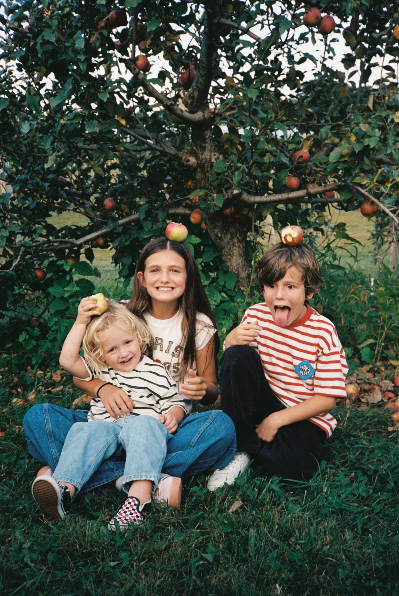 Three children with long and short hair, sitting in front of an apple tree with apples, smiling and playing outside.