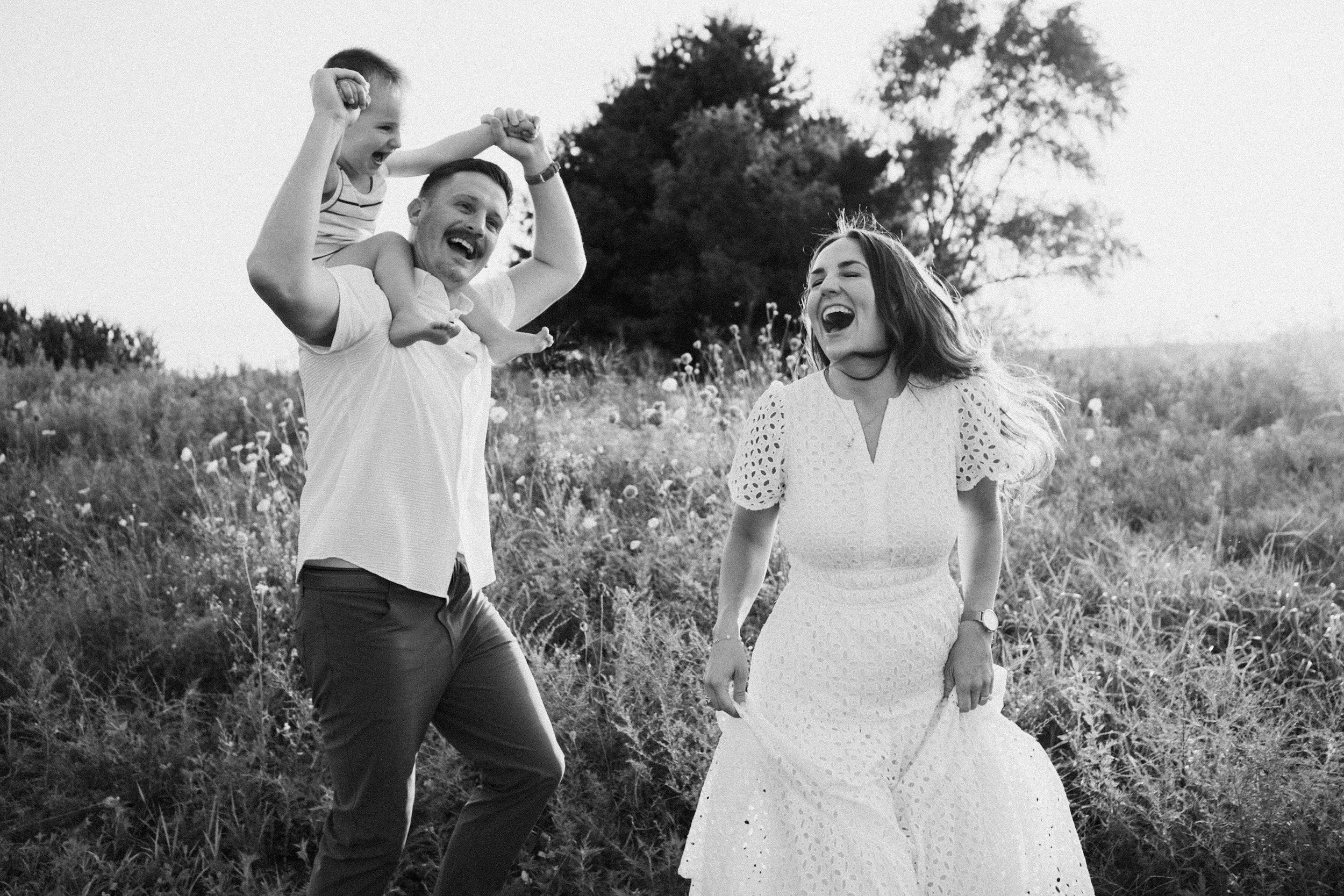 A family of three enjoying outdoors: a man carrying a young boy on his shoulders, and a woman walking beside them, all smiling and laughing in a grassy field with trees in the background.