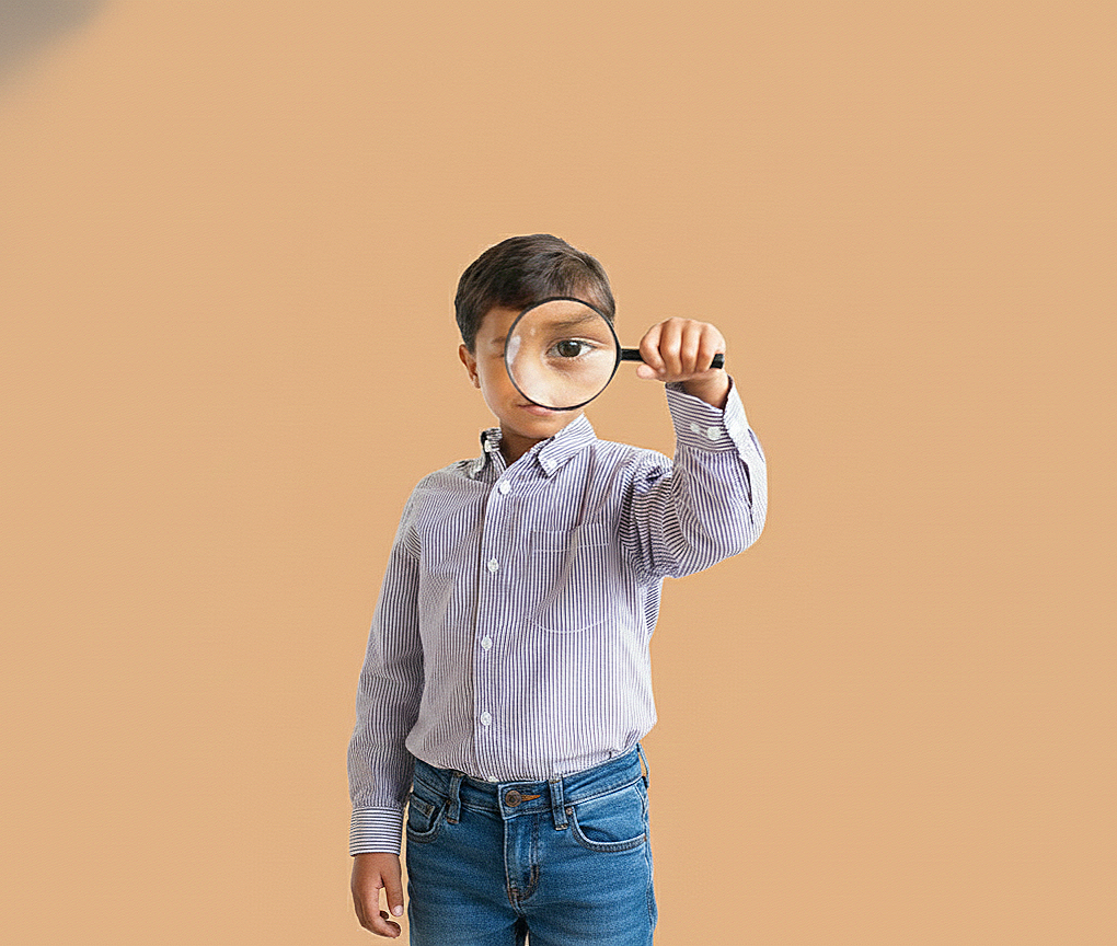 Young boy in a striped shirt holding a magnifying glass up to his eye, standing against a plain beige background.