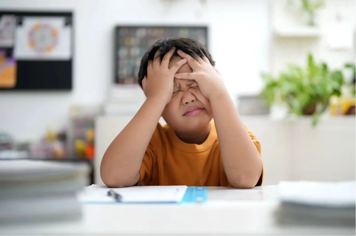 A young boy with dark hair in an orange shirt sitting at a desk with his face scrunched up and hands on his forehead, appearing stressed or upset. The background shows a classroom or study area with blurred objects, including a computer screen and some plants.
