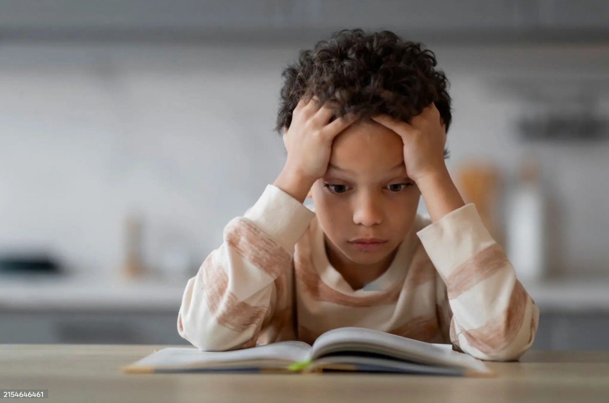 A young boy with curly hair and a distressed expression looks at an open book on a table in a kitchen.