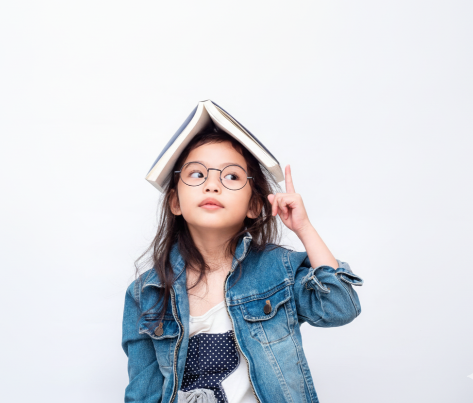 Young girl with glasses and a denim jacket balancing a book on her head, pointing upward with her index finger.