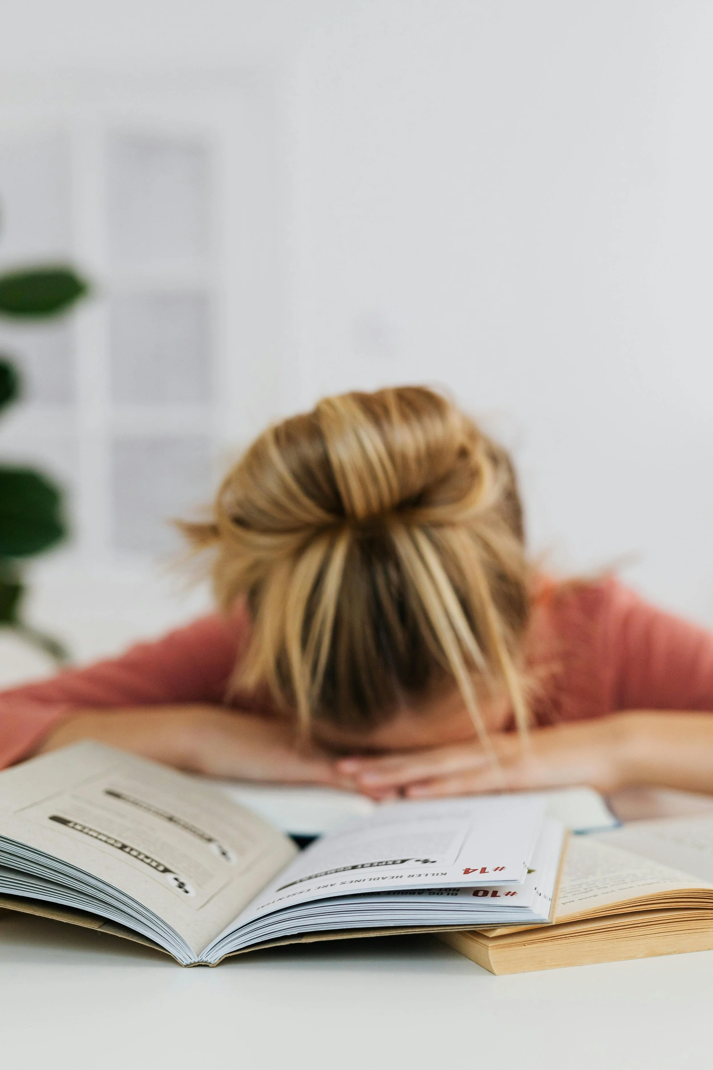 A person with blonde hair resting their head on their arms on a table, surrounded by open books or manuals.