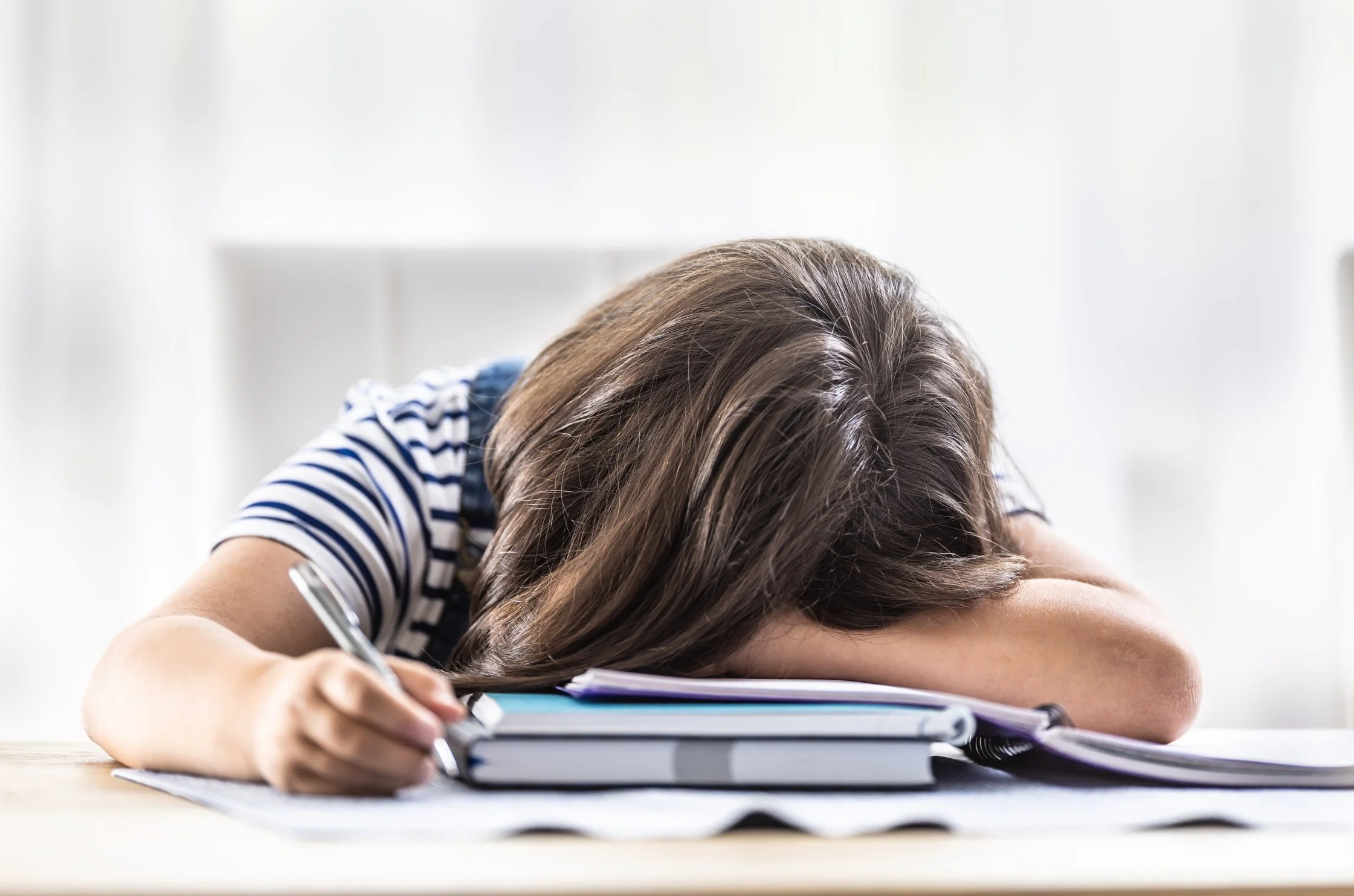 A young girl with brown hair resting her head on the table while doing homework, surrounded by notebooks and papers.