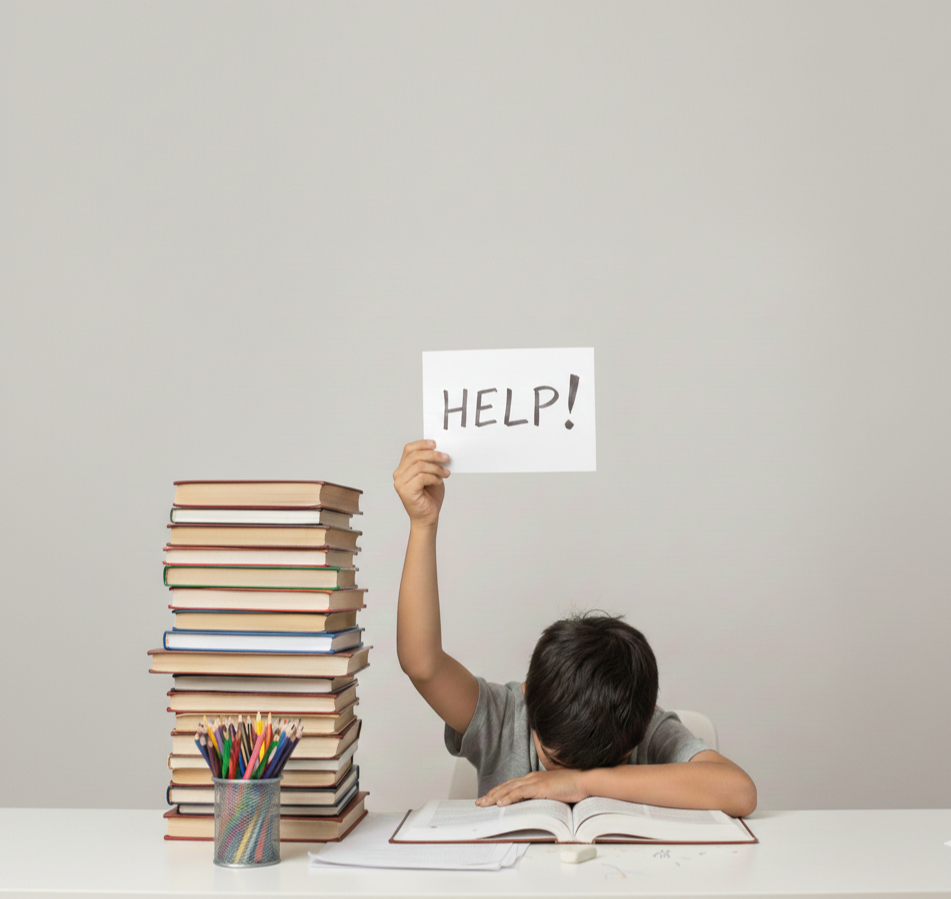 A young boy sitting at a desk with a large stack of books, an open book in front of him, and holding up a sign that says 'HELP!'.