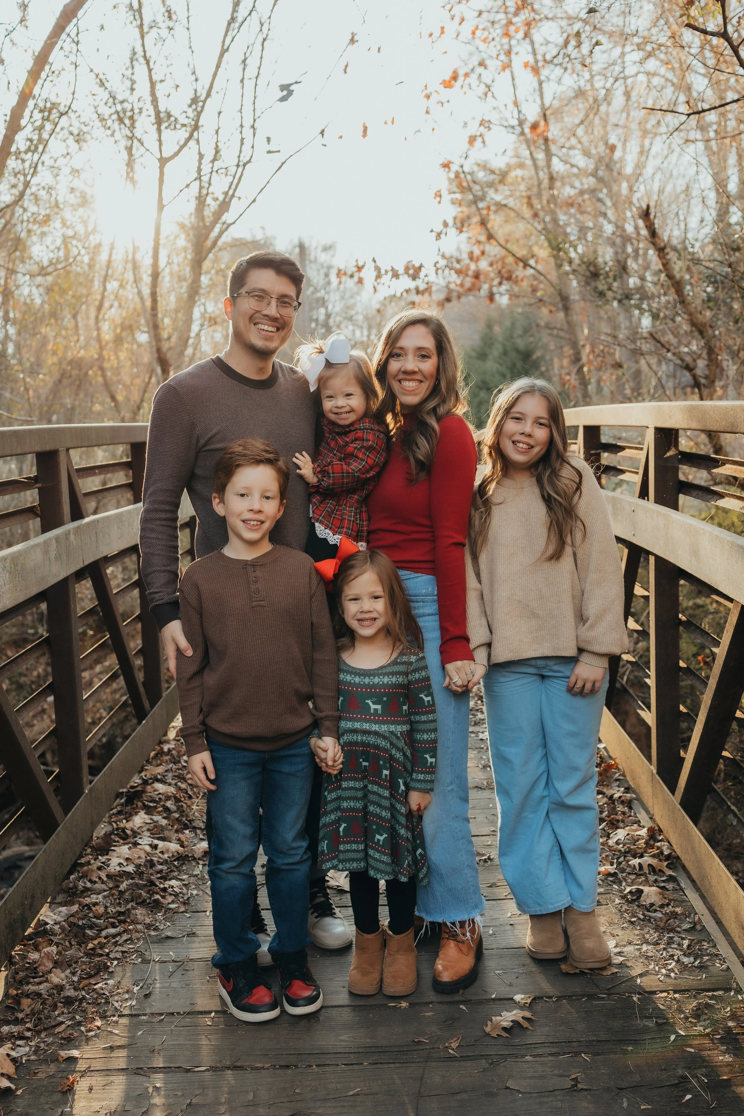 A family of seven standing on a wooden bridge in a wooded area during fall, smiling at the camera.