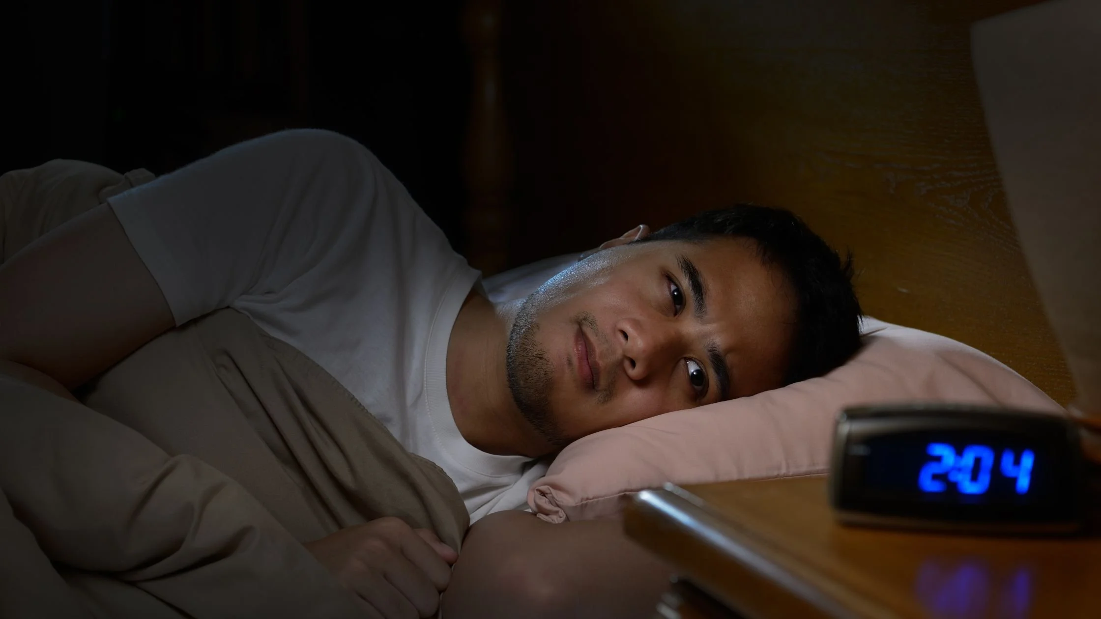 a man laying awake on his side in bed next to a nightstand with an alarm clock that says 2:04 AM