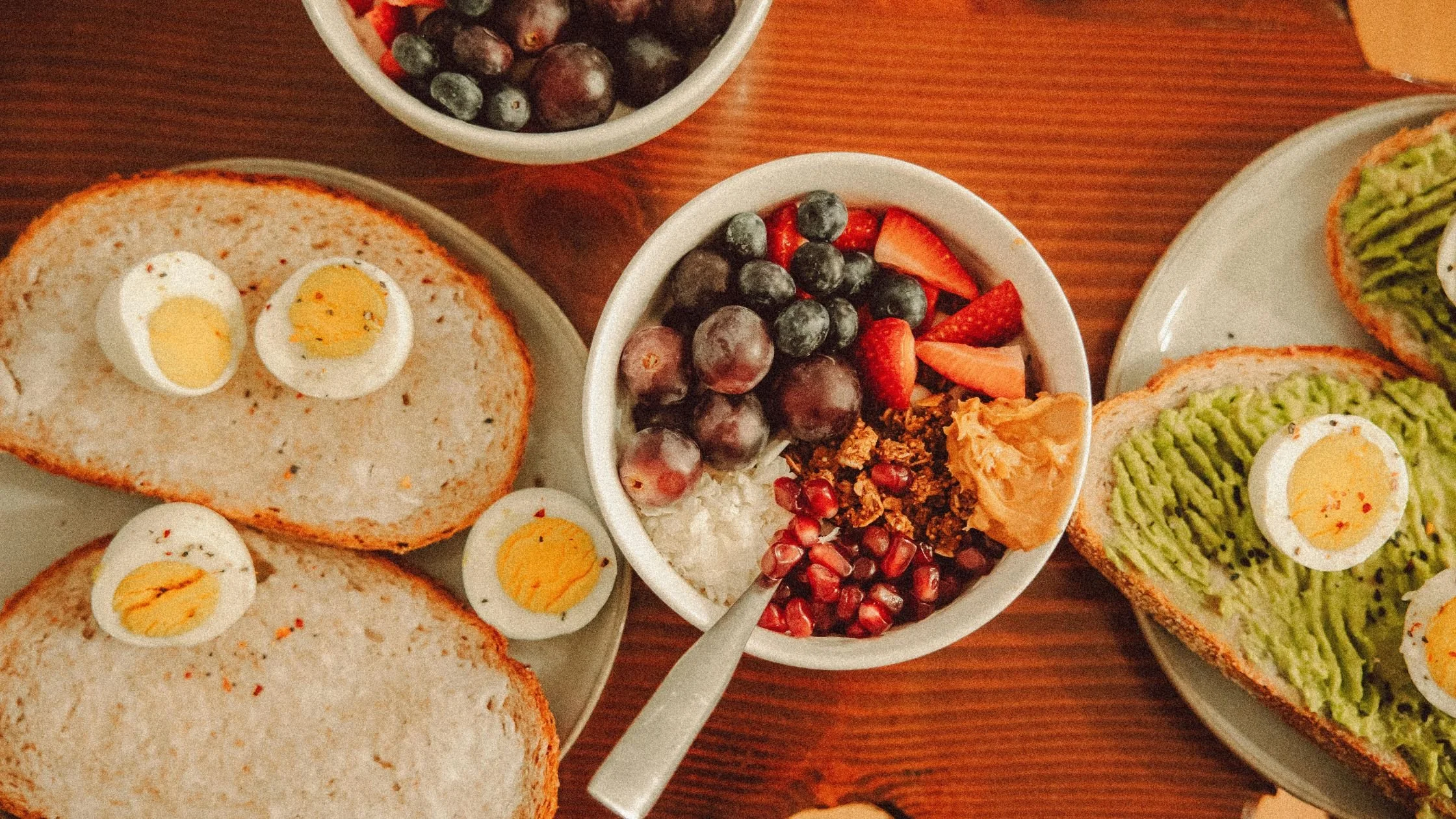 an overhead shot of a brown wooden table with a bowl of fruit and granola, toast and hardboiled eggs, and avocado toast