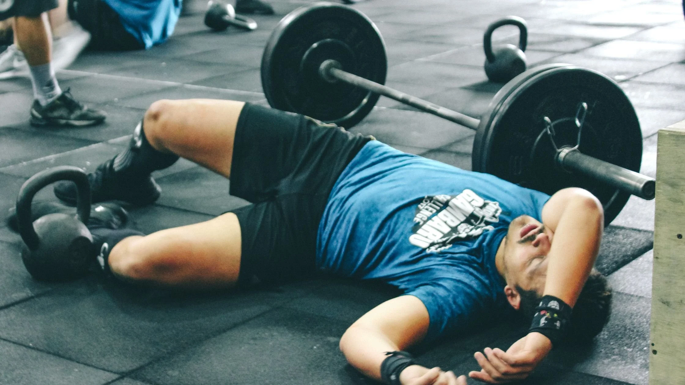 a man sprawled out across the floor of a gym with an arm over his face, looking exhausted next to a barbell and a kettlebell