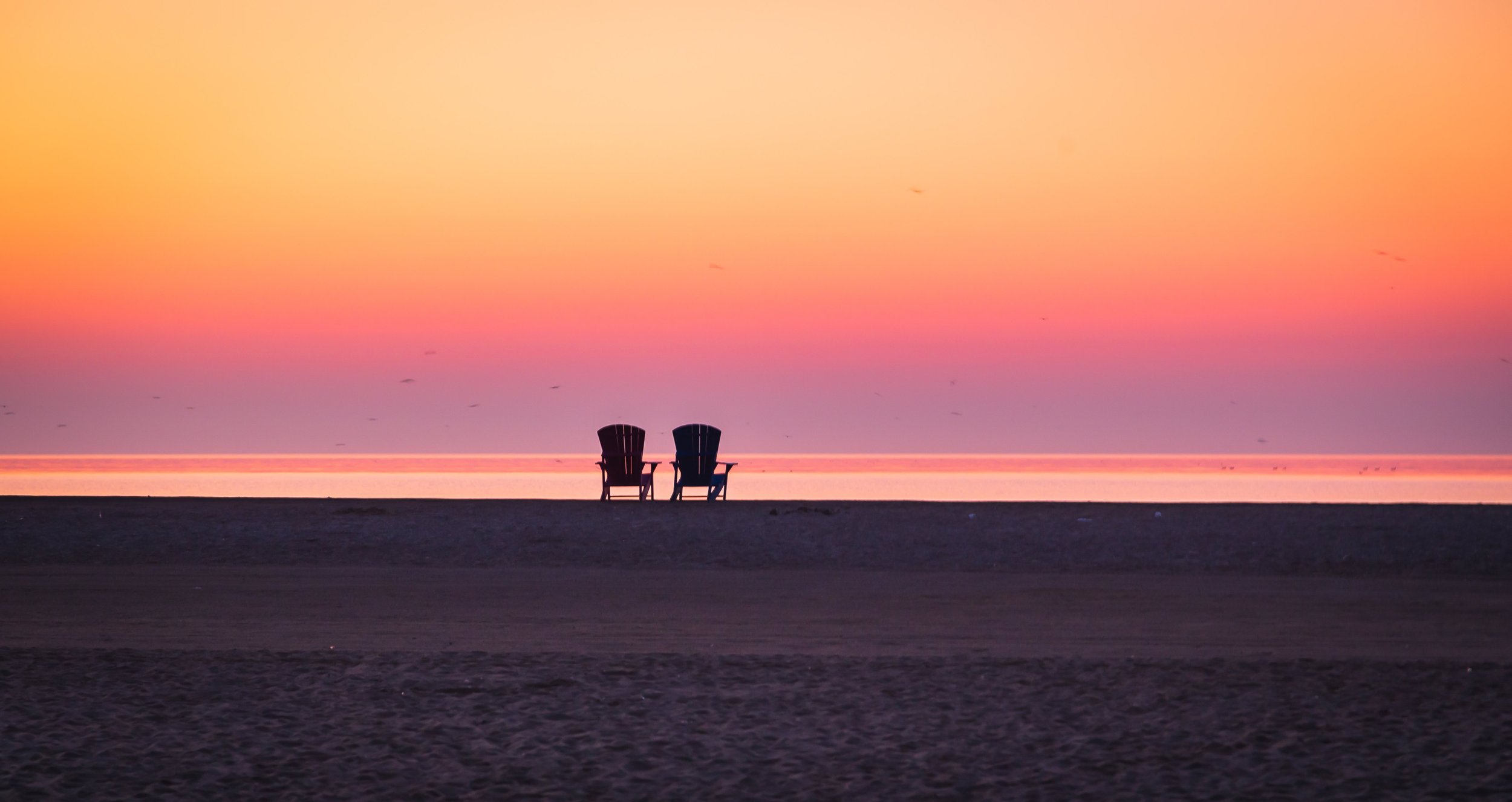 Two empty chairs silhouetted against a warm orange and pink sunset over the water in Toronto, Canada.