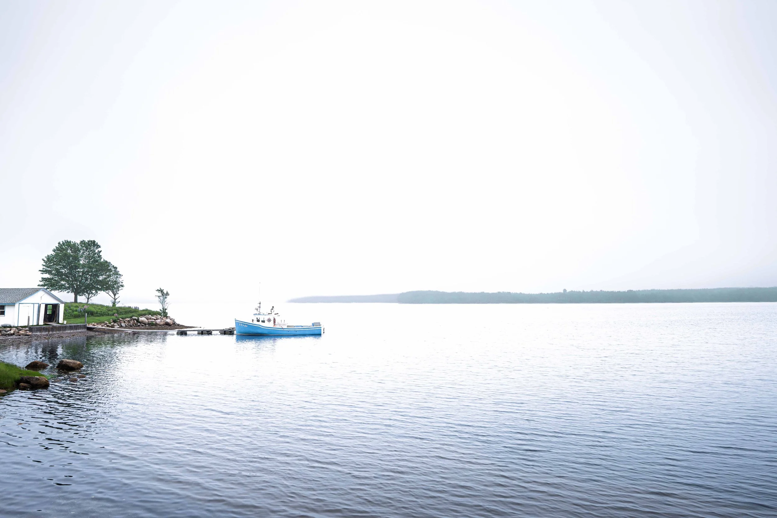 Small blue boat near a quiet shoreline surrounded by expansive water and pale sky in Cape Breton, Canada.
