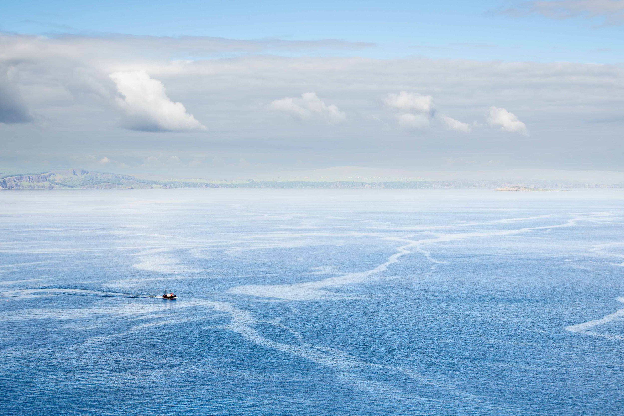 Small solitary boat crossing an expansive blue seascape beneath soft clouds in Scotland.