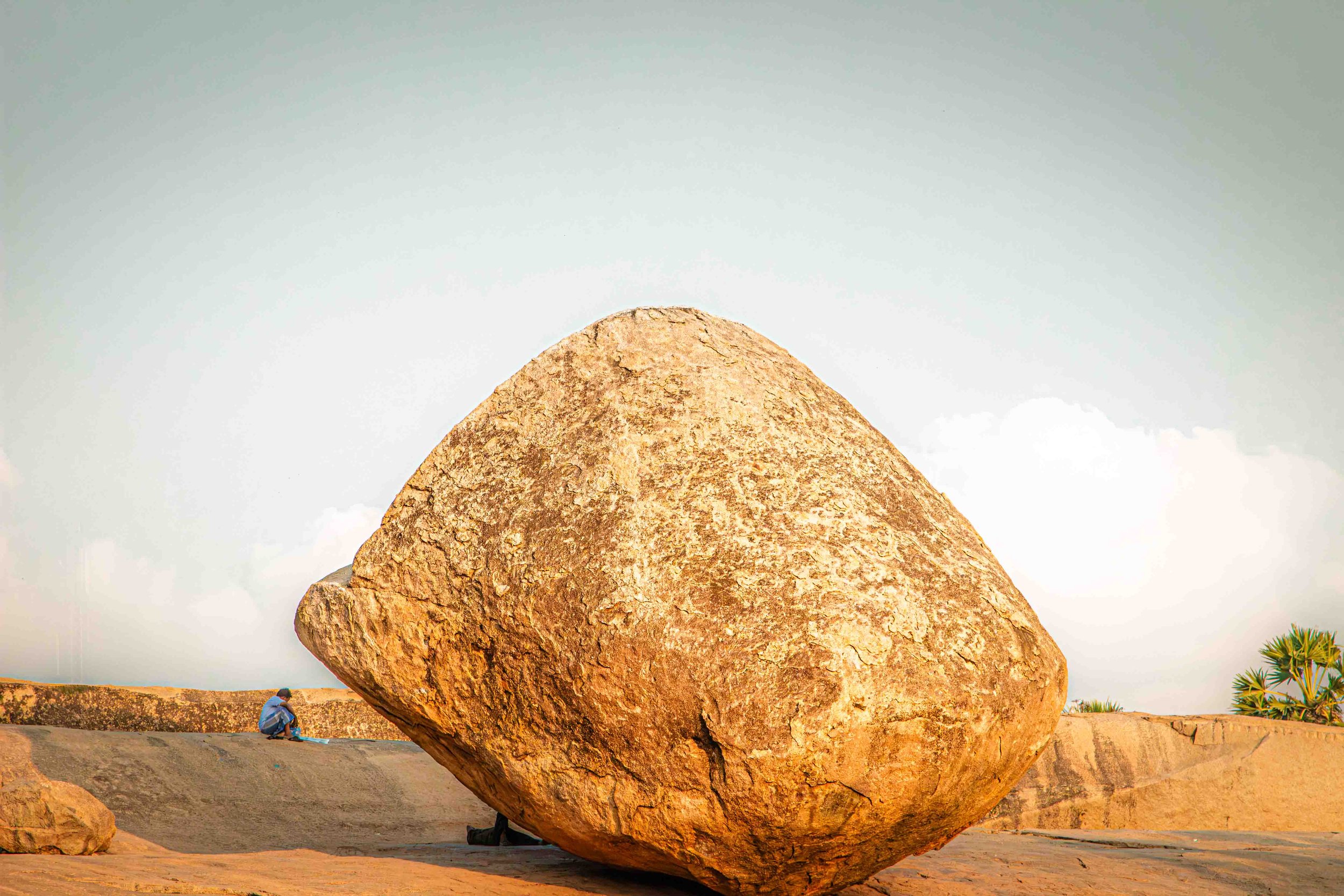 Large sunlit boulder beside a solitary seated person reading in Mahabalipuram, Tamil Nadu, India.