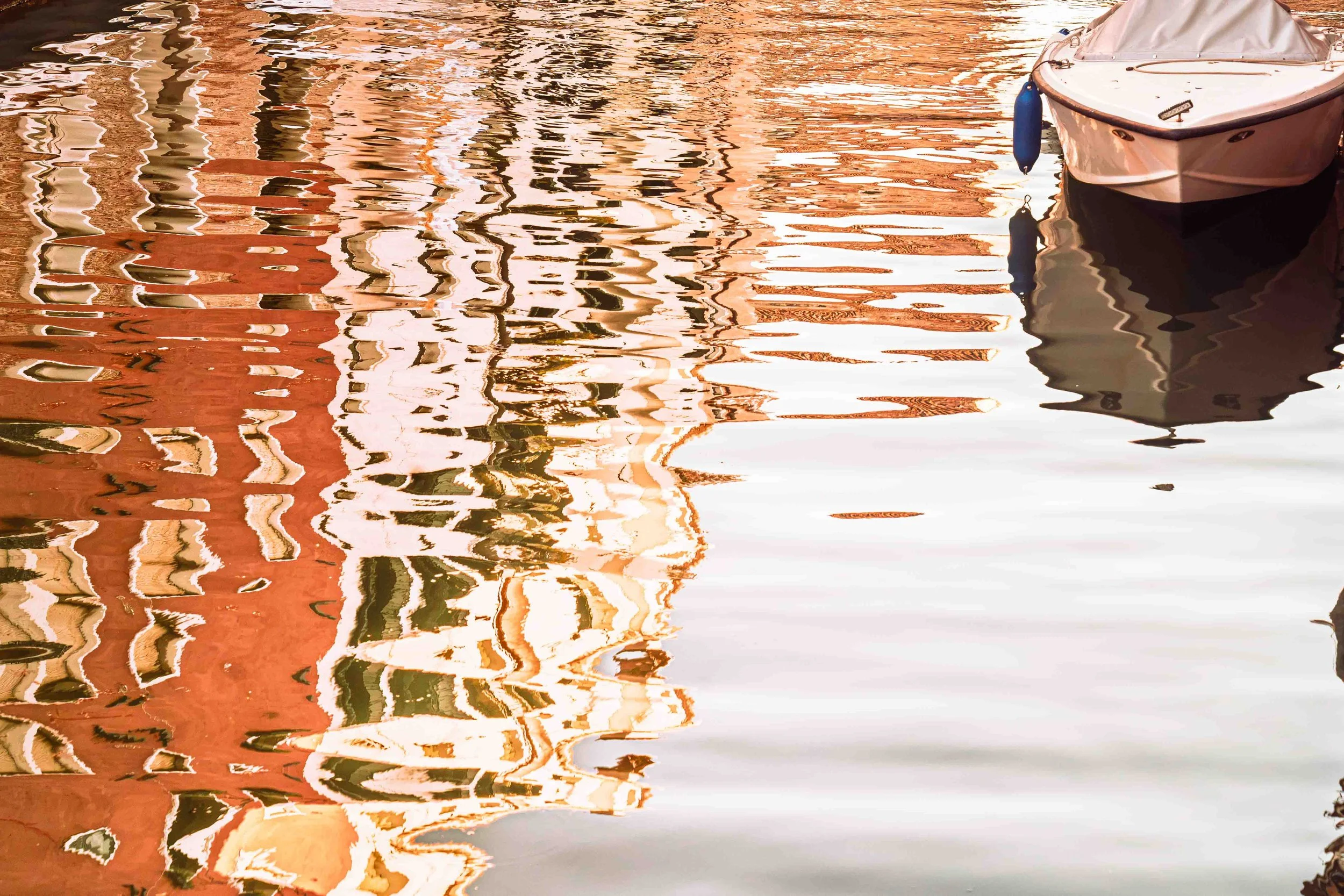 Abstract orange, white and dark reflections of Venetian architecture rippling across water beside a boat in Venice, Italy.