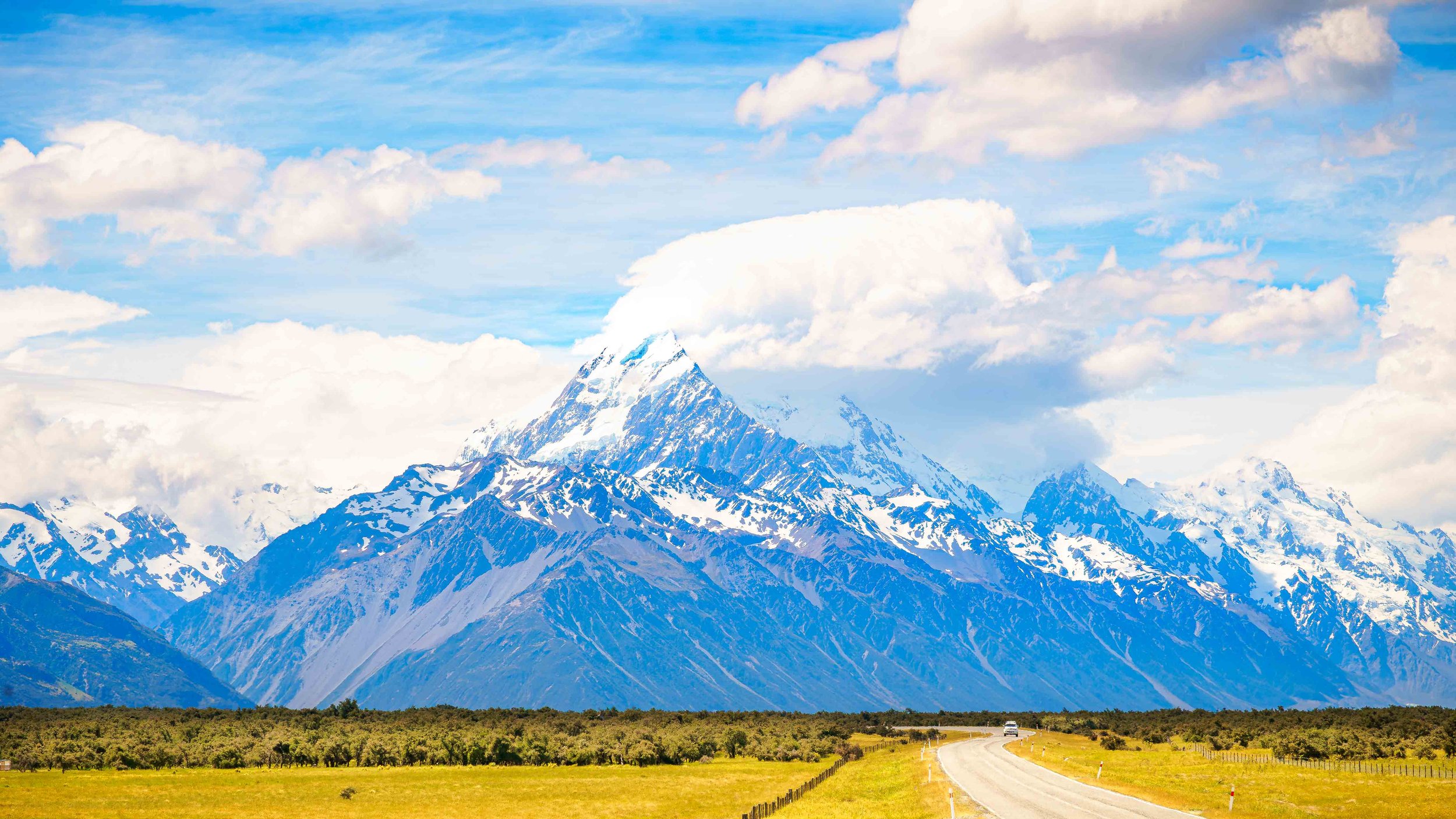 Road leading toward snow-covered Aoraki Mount Cook beneath blue sky and clouds in New Zealand.