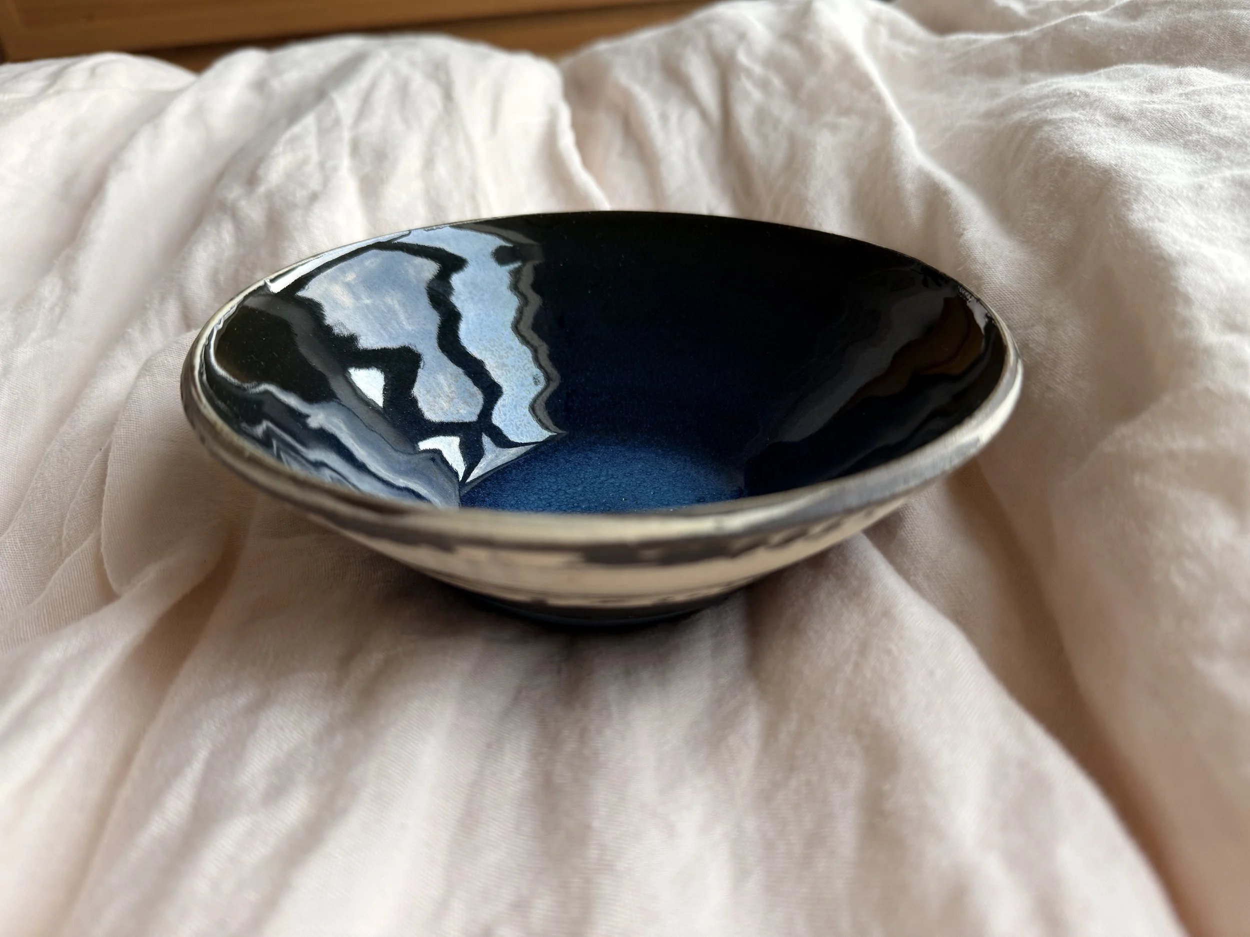 A dark blue ceramic bowl with a glossy finish, placed on a soft, white fabric background.
