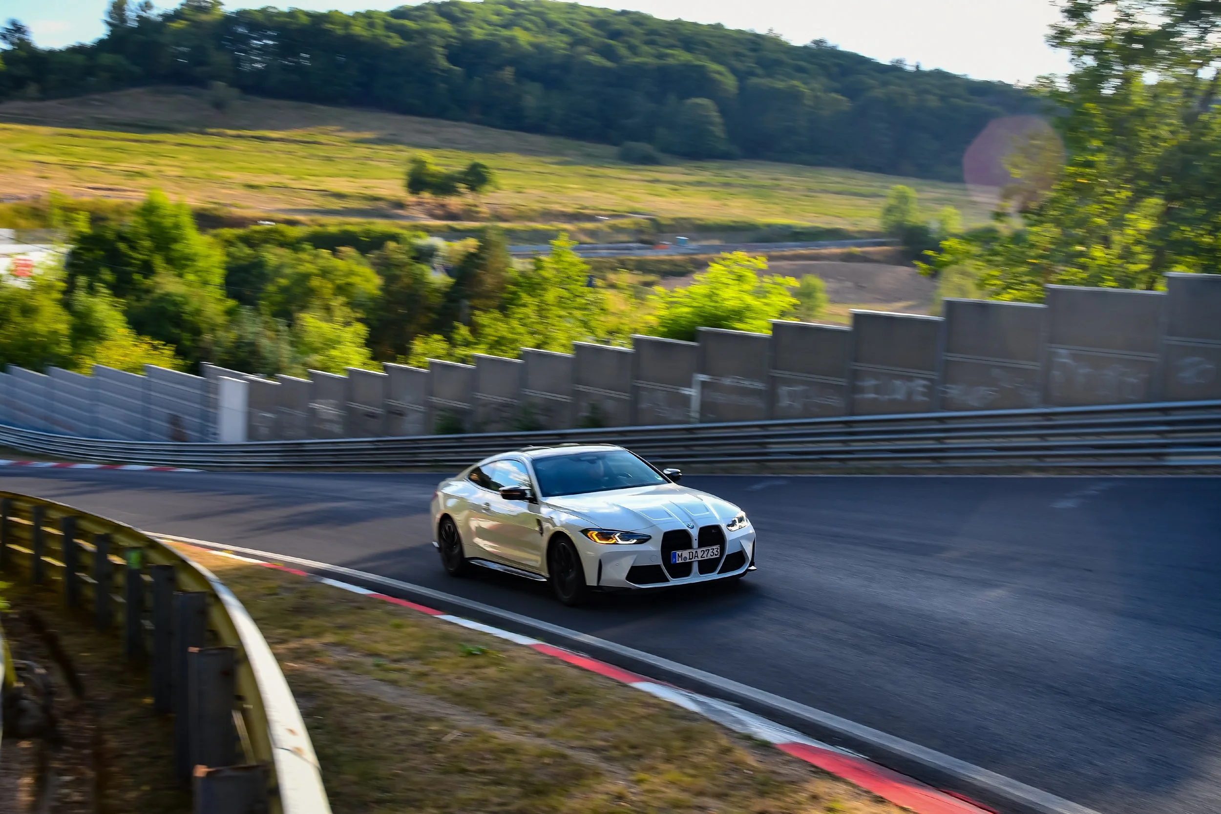 White sports car driving on a racetrack with trees and a barrier in the background.