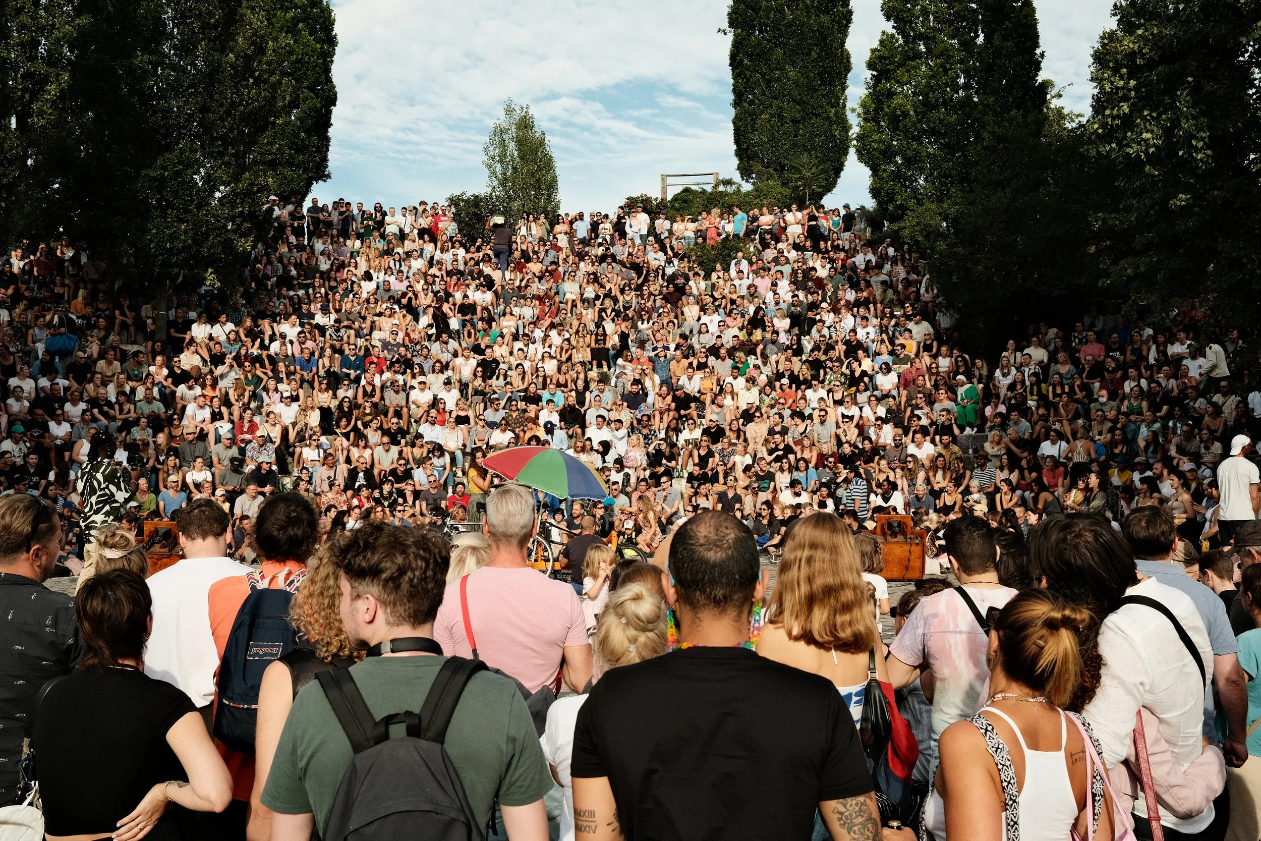 Large crowd gathered in an outdoor amphitheater, surrounded by trees, with a mix of seated and standing attendees.