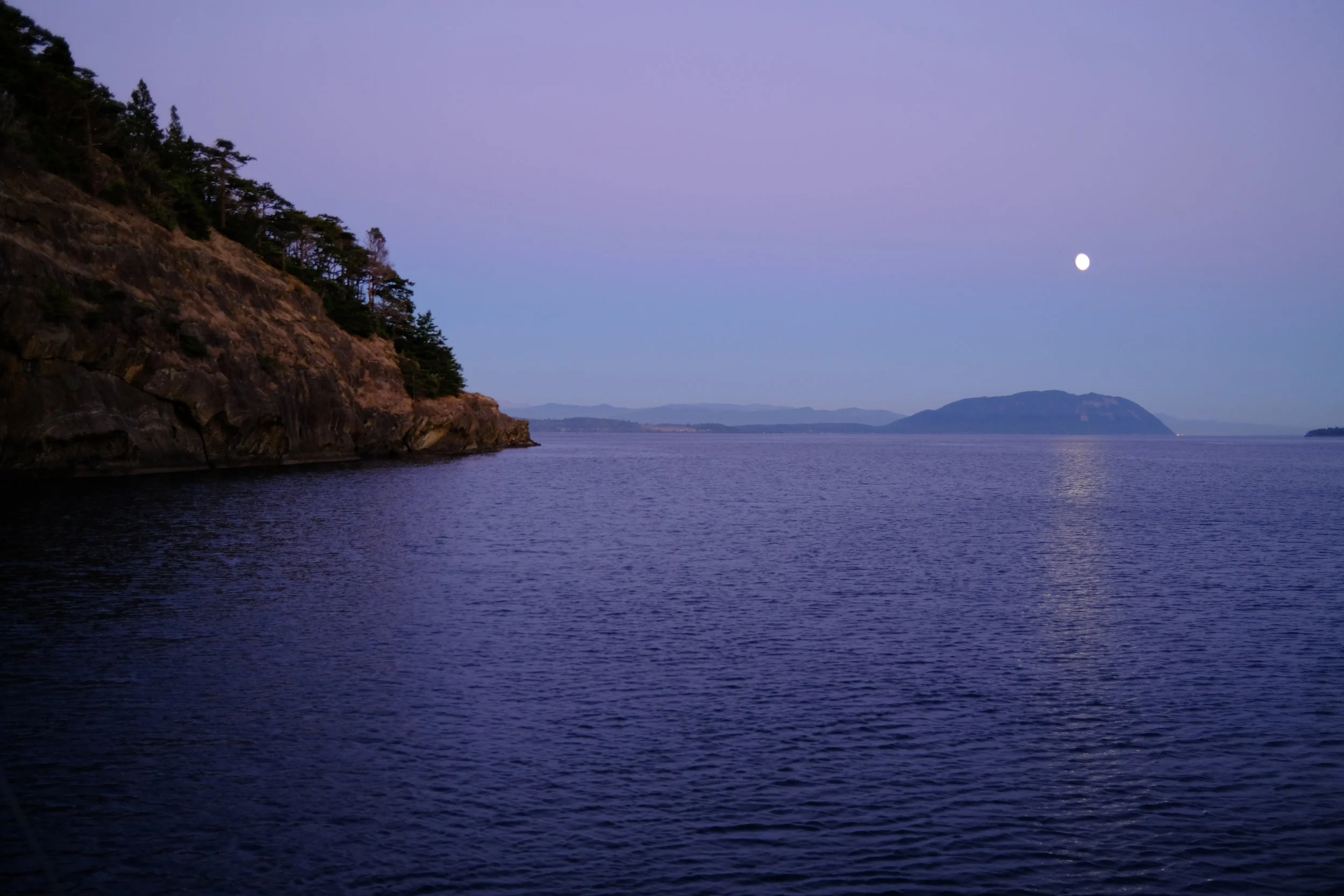 Coastal landscape with cliffs, ocean, moon, and distant mountains at twilight.