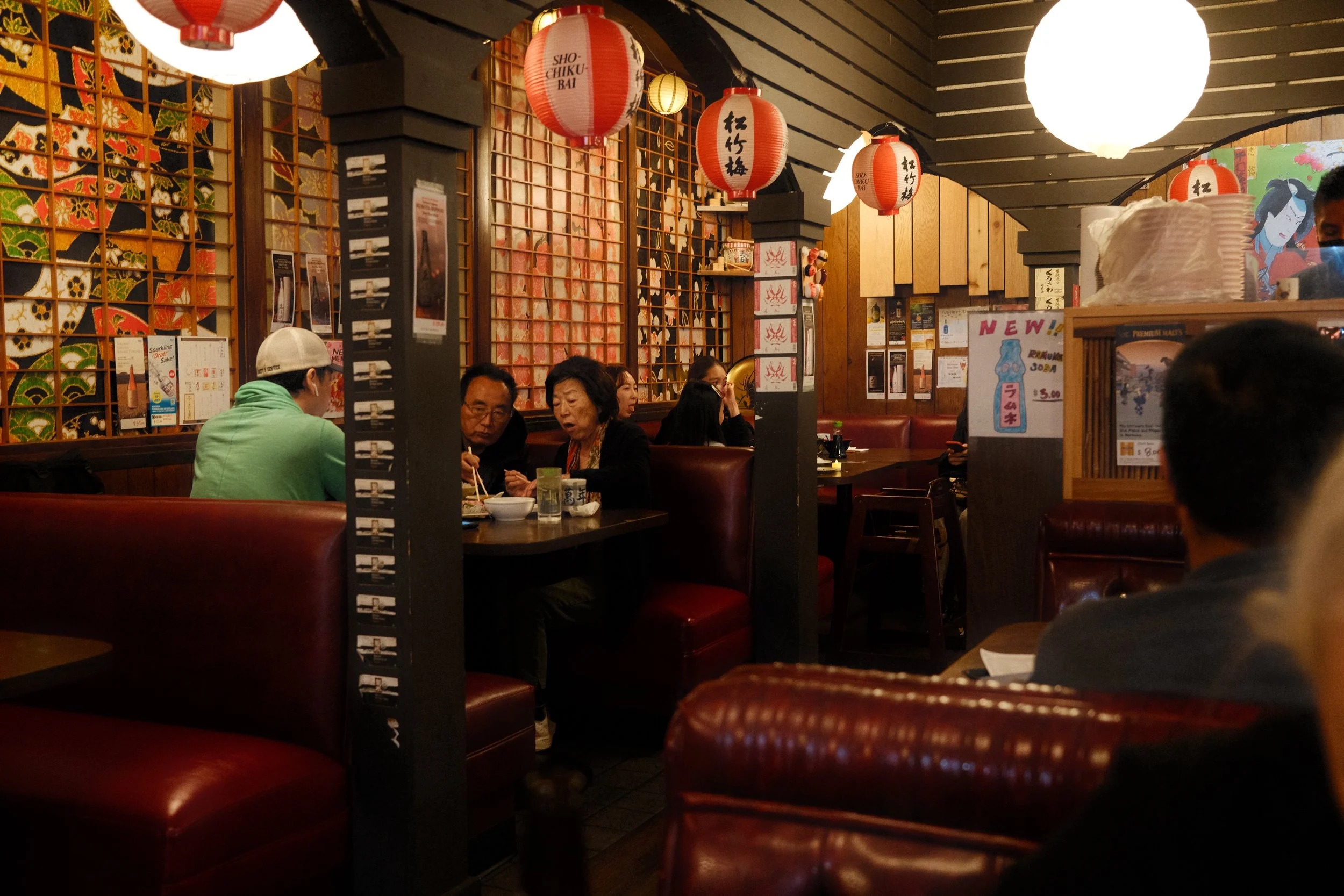 Interior of a Japanese restaurant with red booths and lanterns, featuring diners enjoying meals. Walls decorated with colorful artwork and Japanese writing.