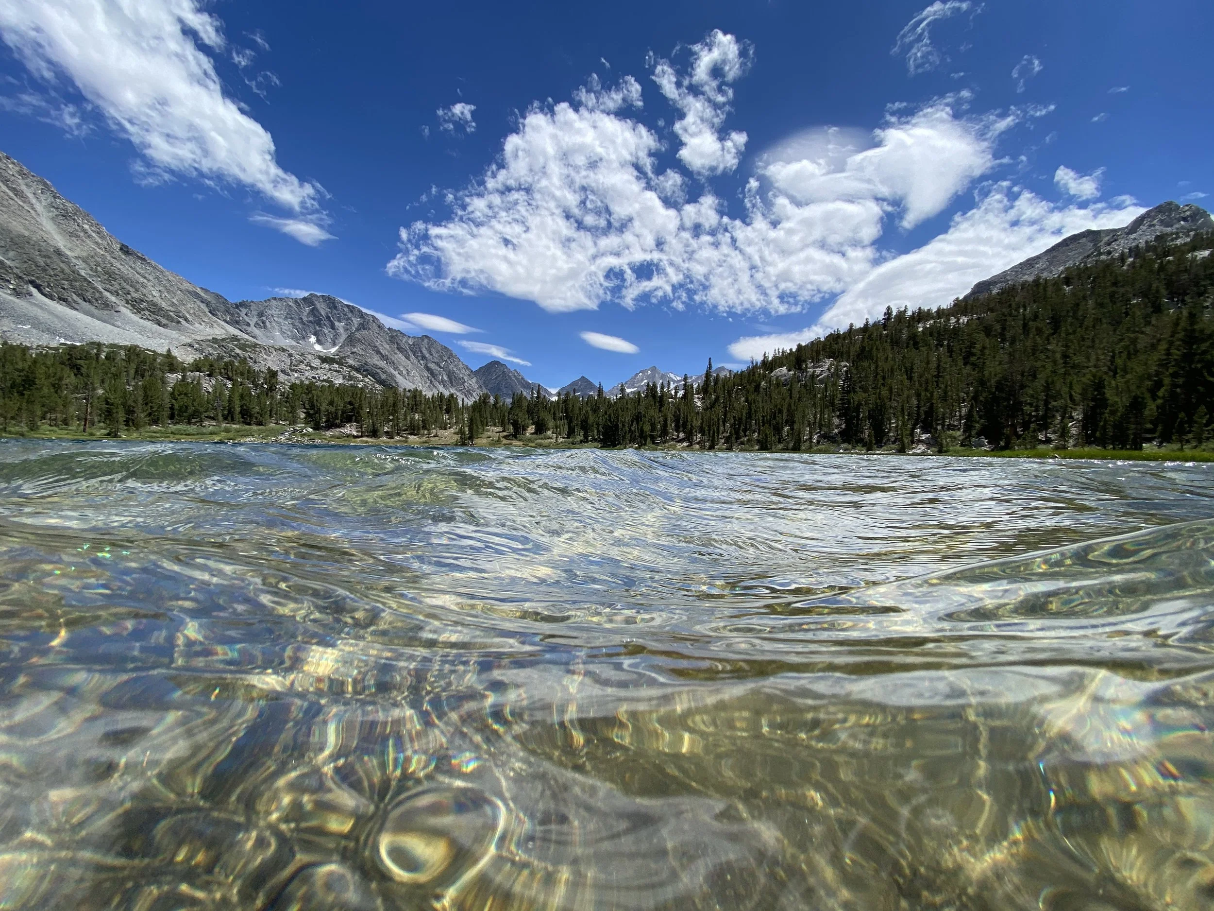 Mountain lake with clear water, surrounded by pine trees and rocky mountains under a blue sky with clouds.