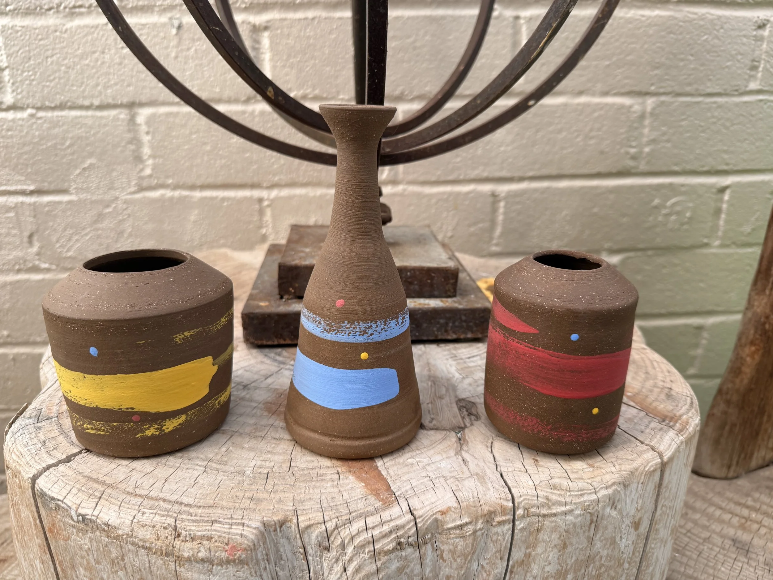 Three handcrafted clay vases with colorful painted designs, set on a wooden surface against a white brick background. The vases feature yellow, blue, and red accents.