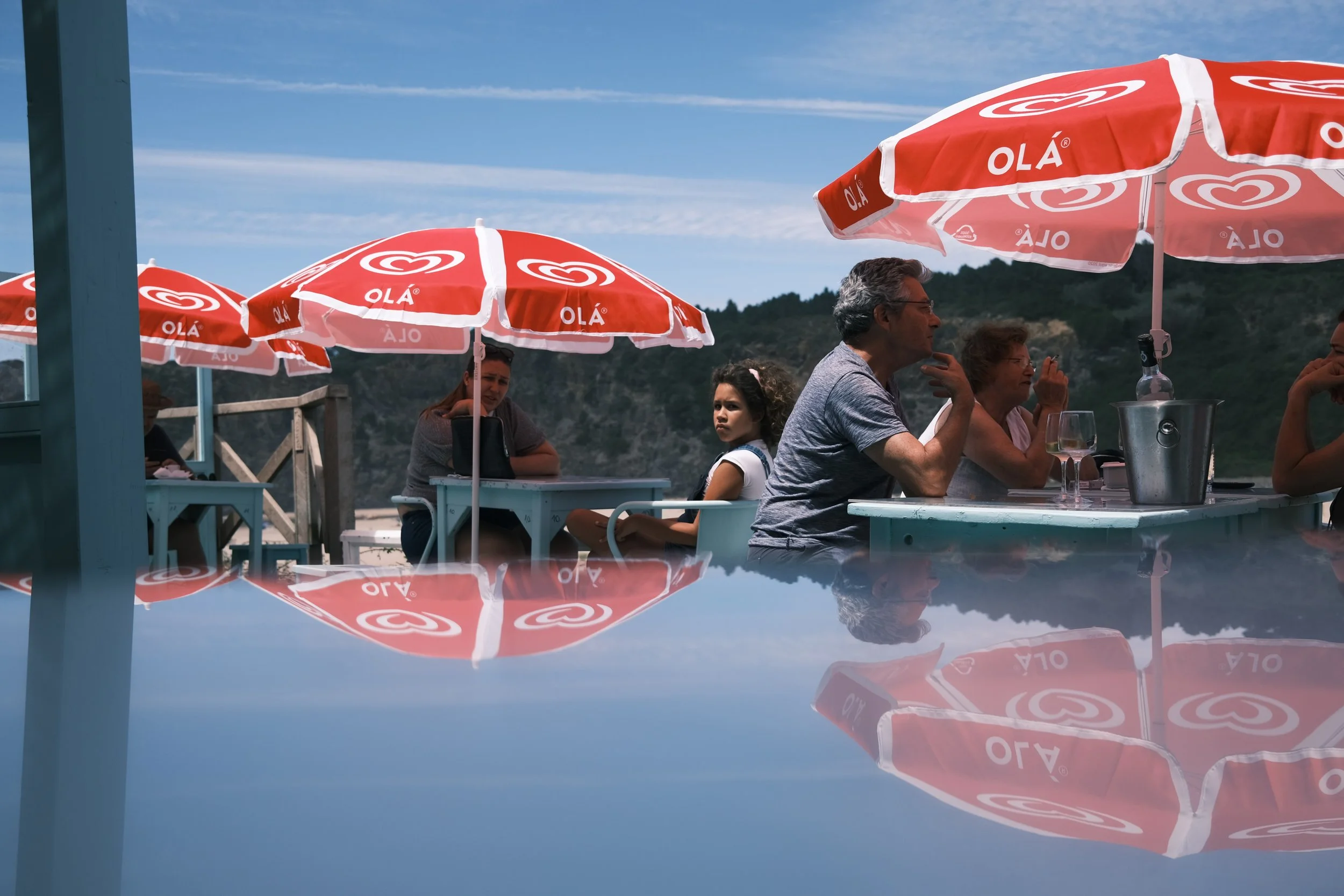 Outdoor café scene with people seated under red and white umbrellas, mountains in the background, and a reflective surface in the foreground.