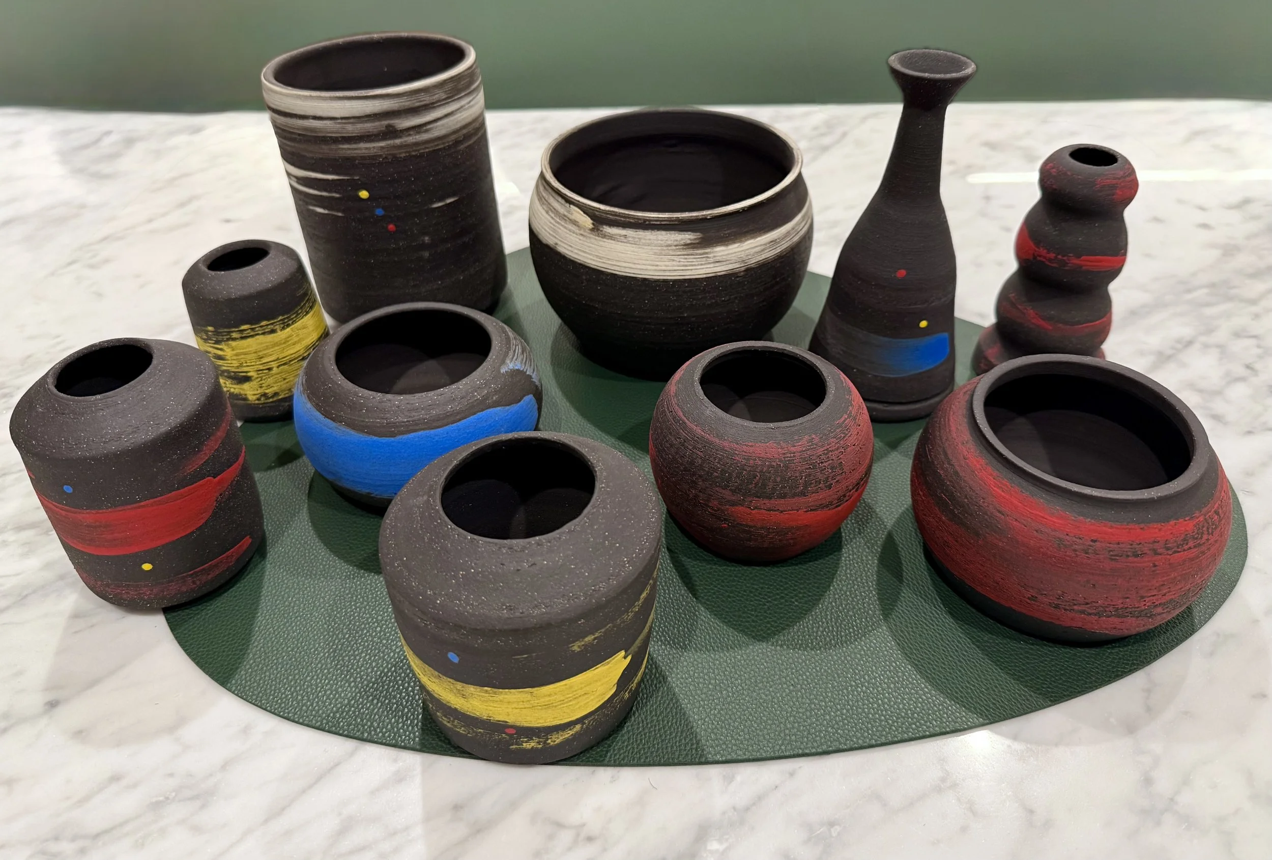 A collection of black ceramic vases and pots with colorful painted accents in red, yellow, blue, and white, arranged on a green surface.